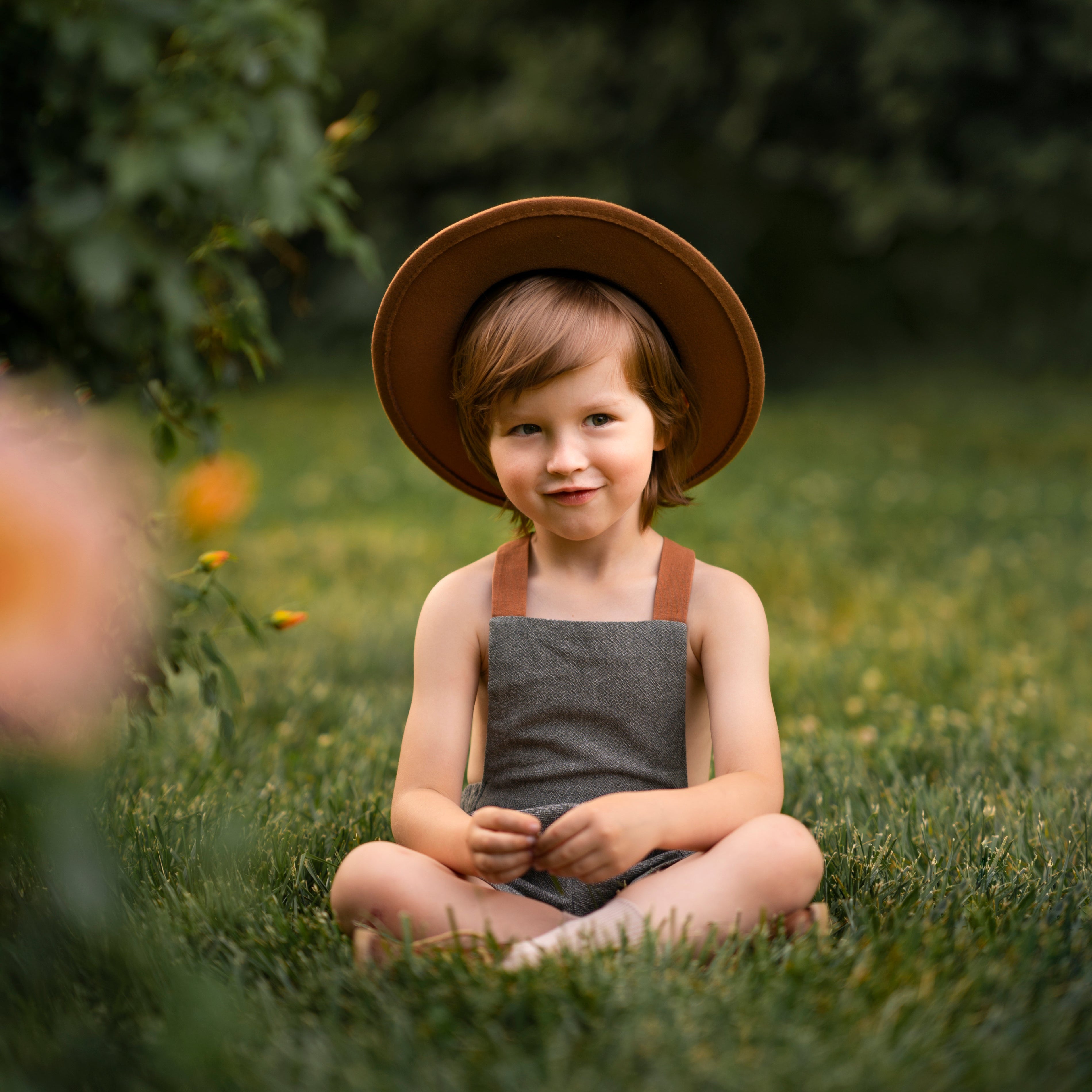 Little boy in green overalls and a hat sitting with cross legs on grass