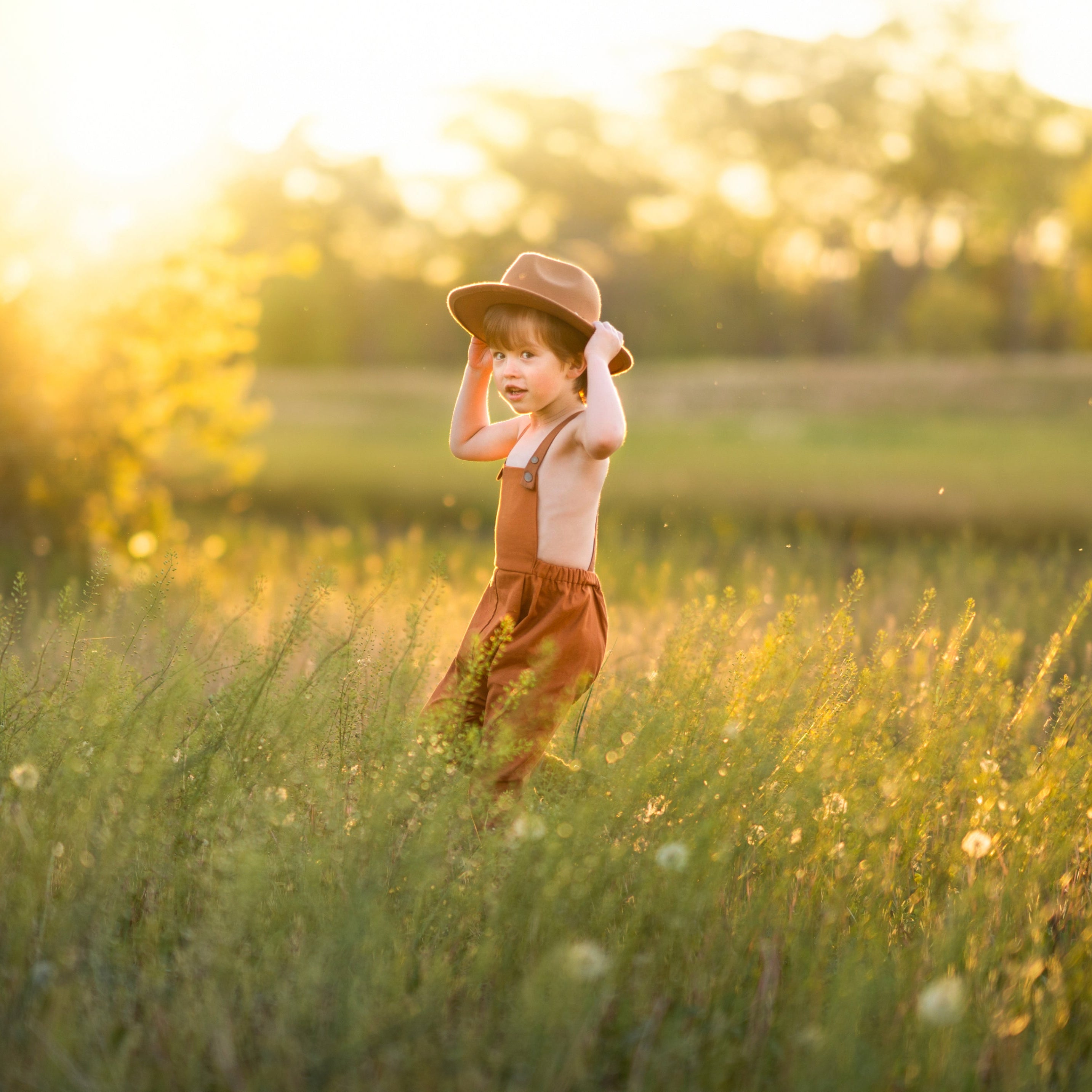 little boy in a field in orange overalls with a hat smiling