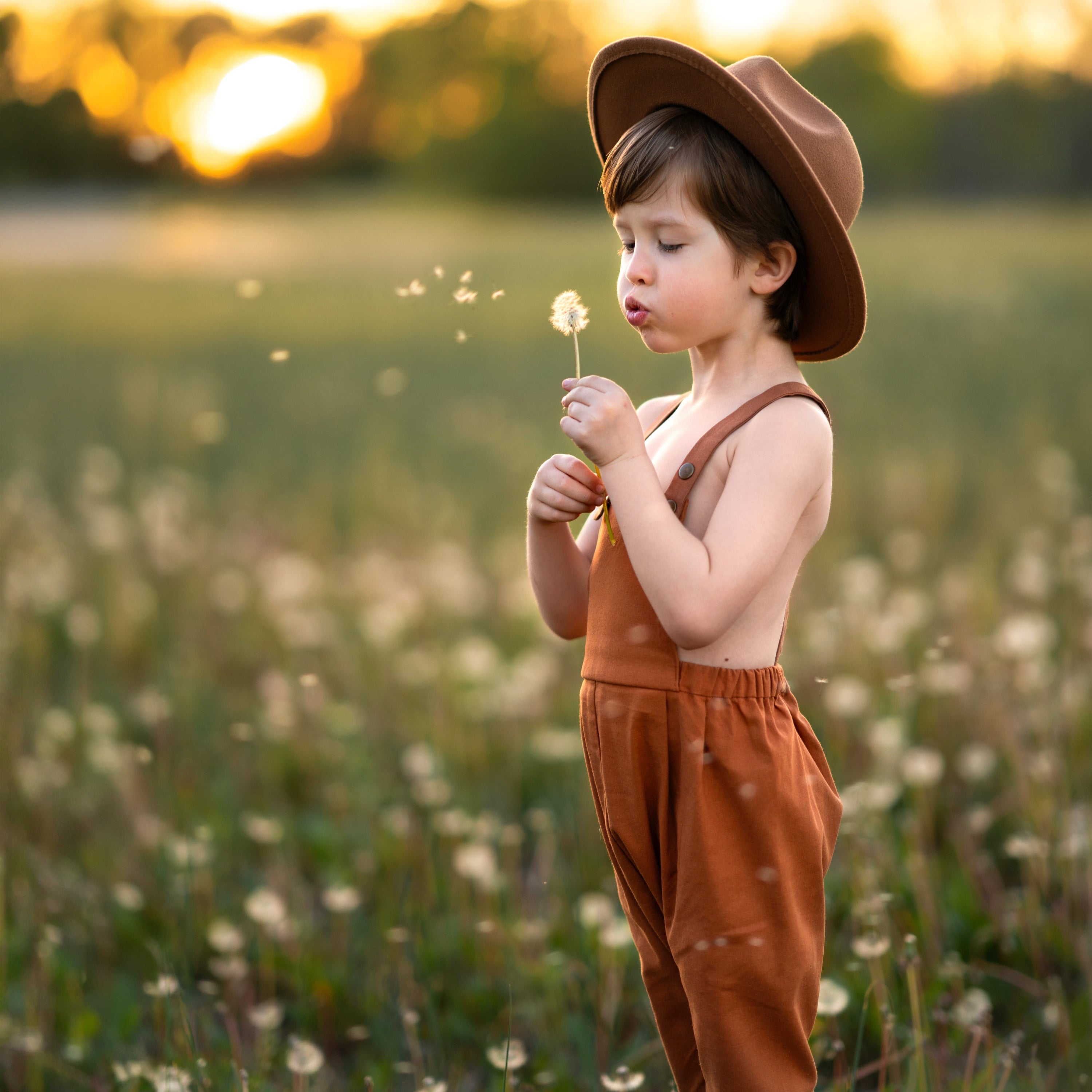 little boy in a field in orange overalls blowing dandelions