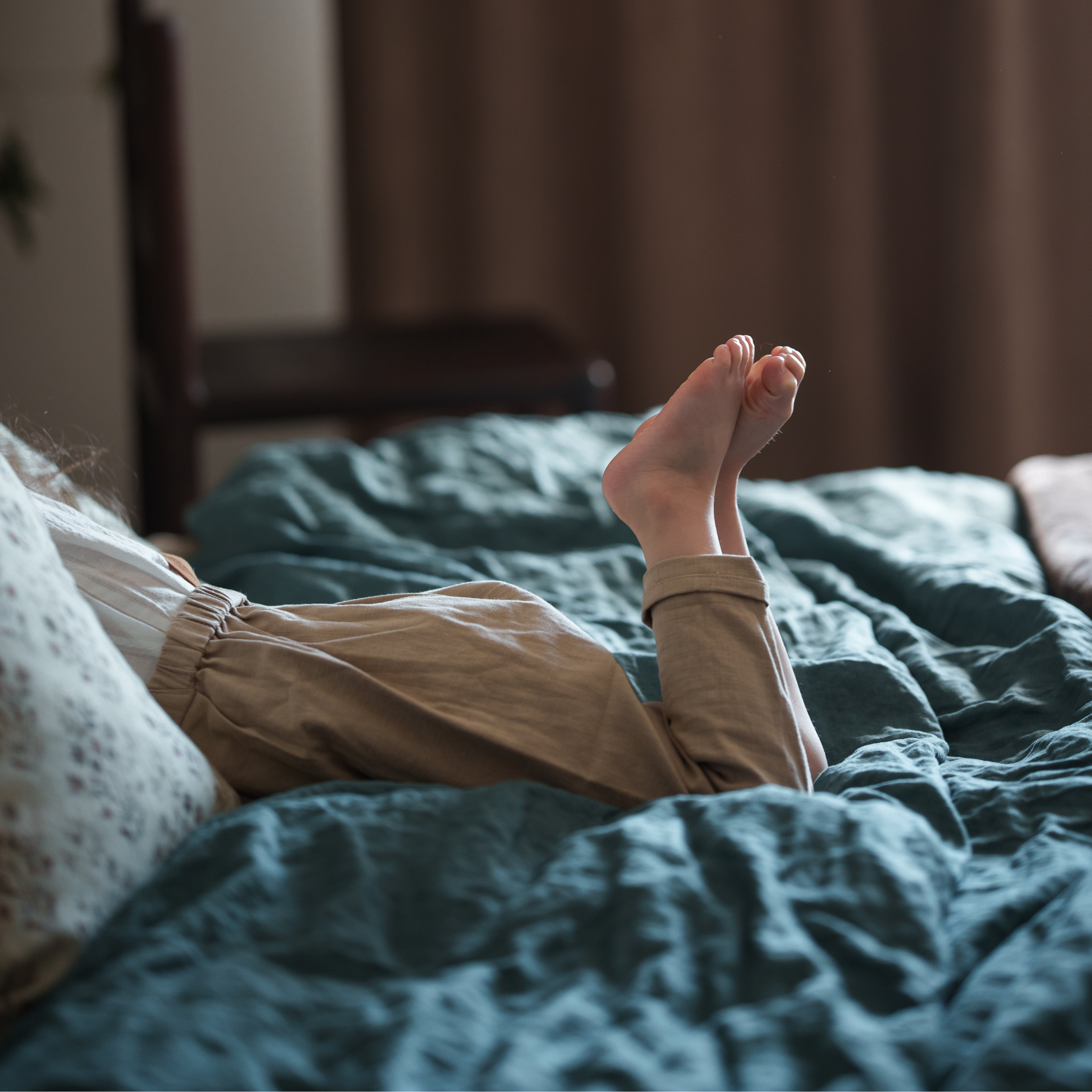 Person lying on a bed with feet raised, wearing beige pants and white socks, in a softly lit room.