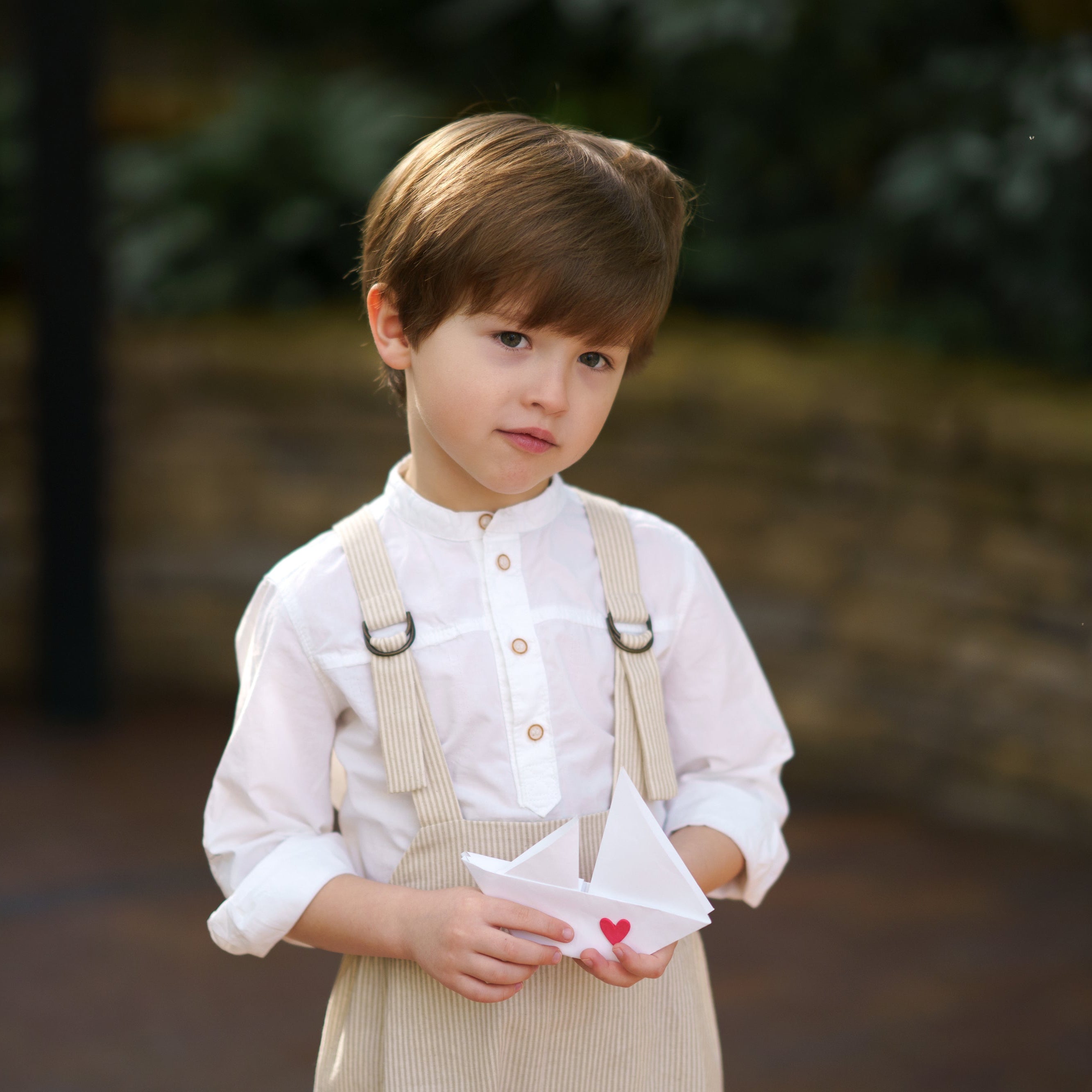 Little boy showcasing details of the Grow-With-Me Romper - Beige Stripes, highlighting the fully adjustable straps for a customizable fit, the classic timeless beige stripes pattern, and the soft, breathable fabric that ensures comfort throughout the day, representing stylish versatility in sustainable children's attire.