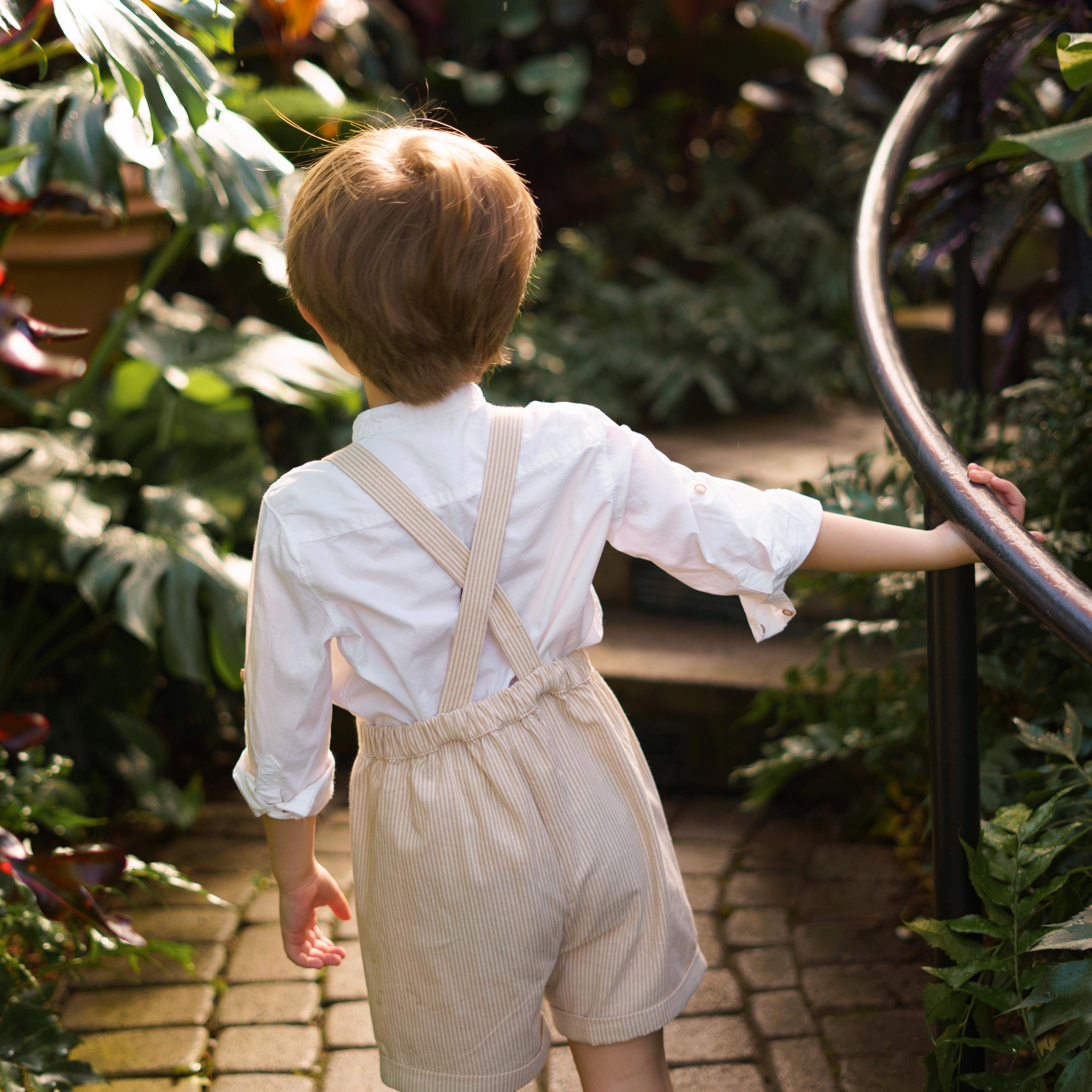 The back of a little boy wearing a beige stripes Grow-With-Me Romper, walking joyfully in a green house, highlighting the adjustable straps and comfortable fit.