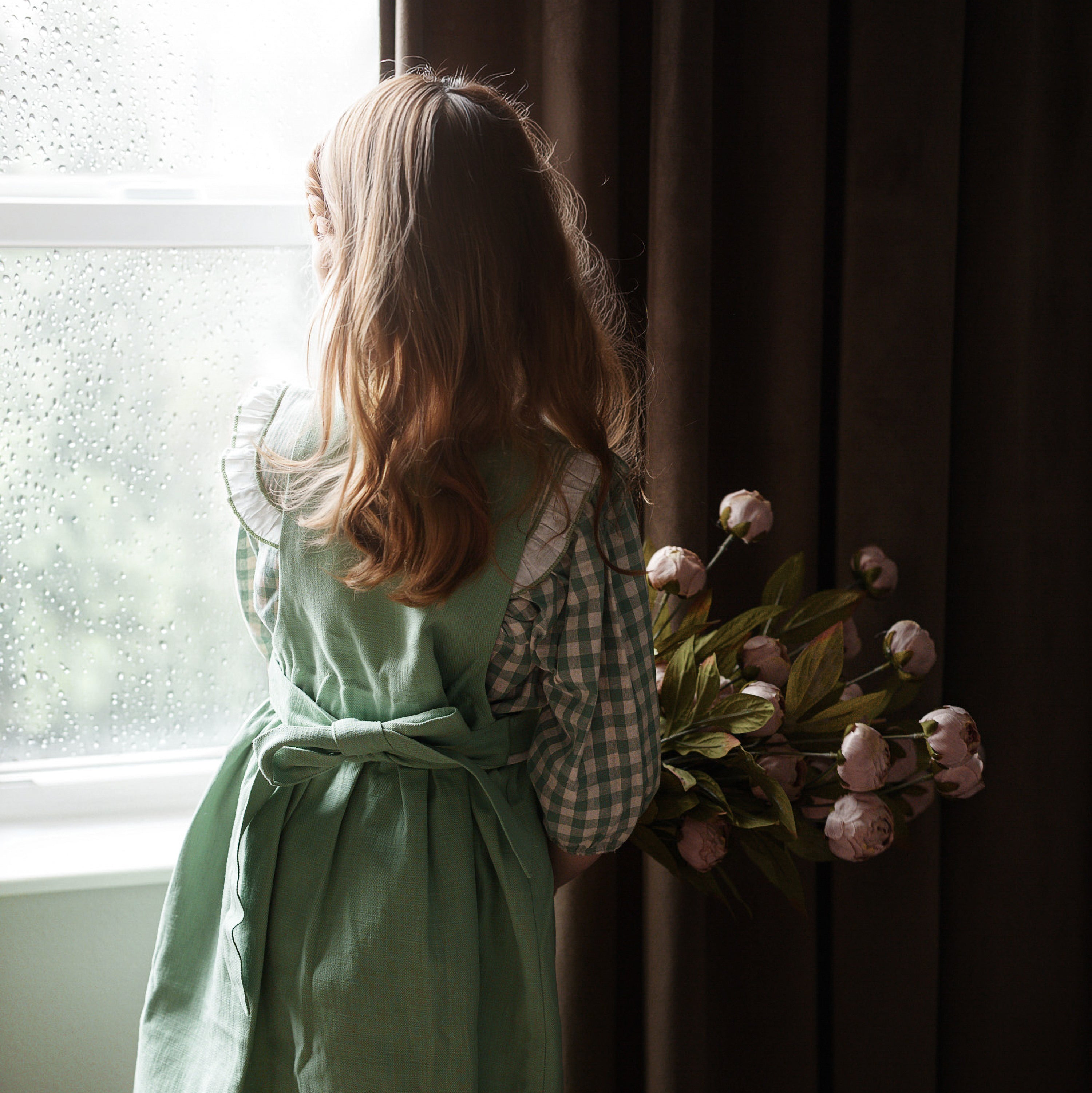 Child in a green dress standing by a window with a bouquet of flowers.