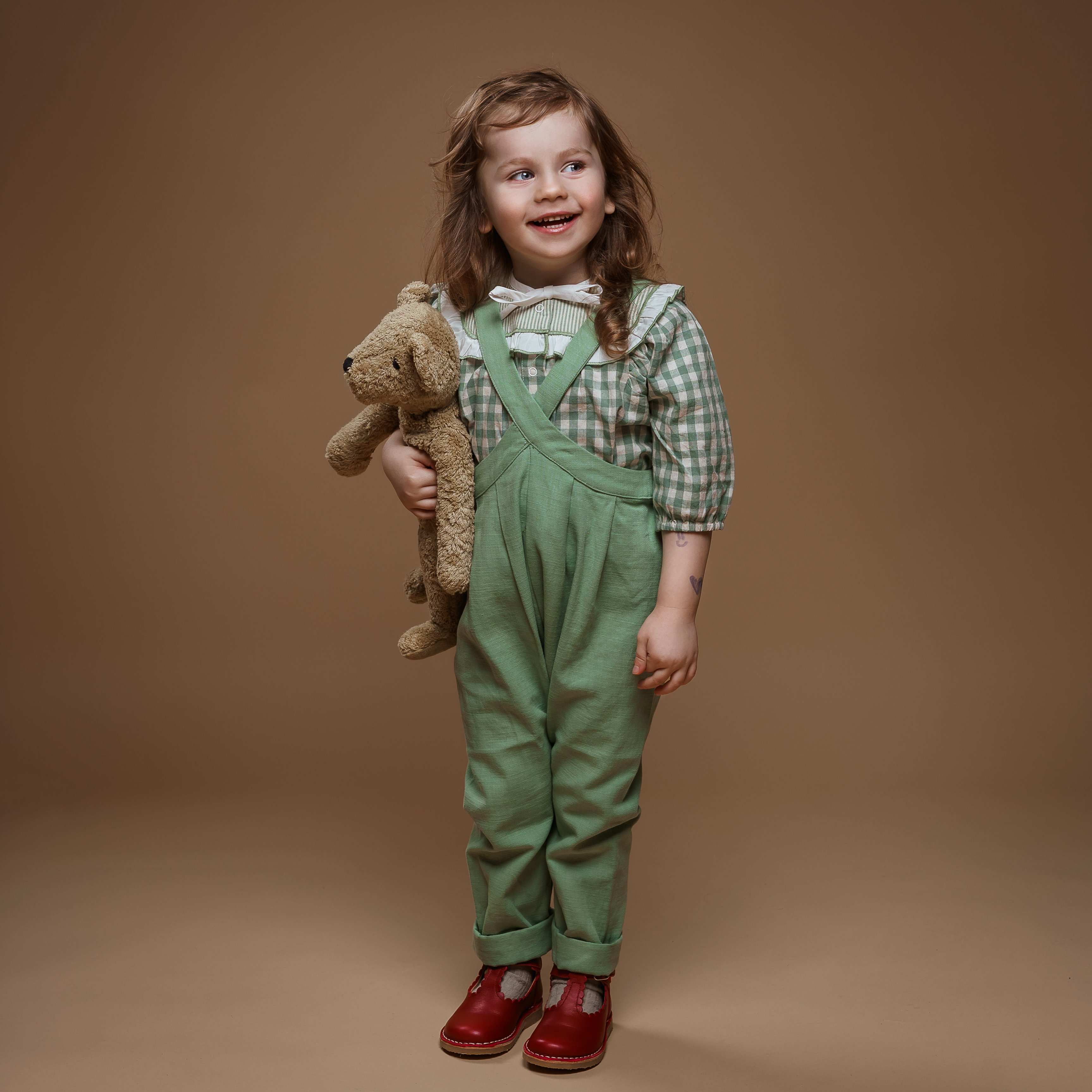 Child wearing green overalls and holding a teddy bear against a brown background