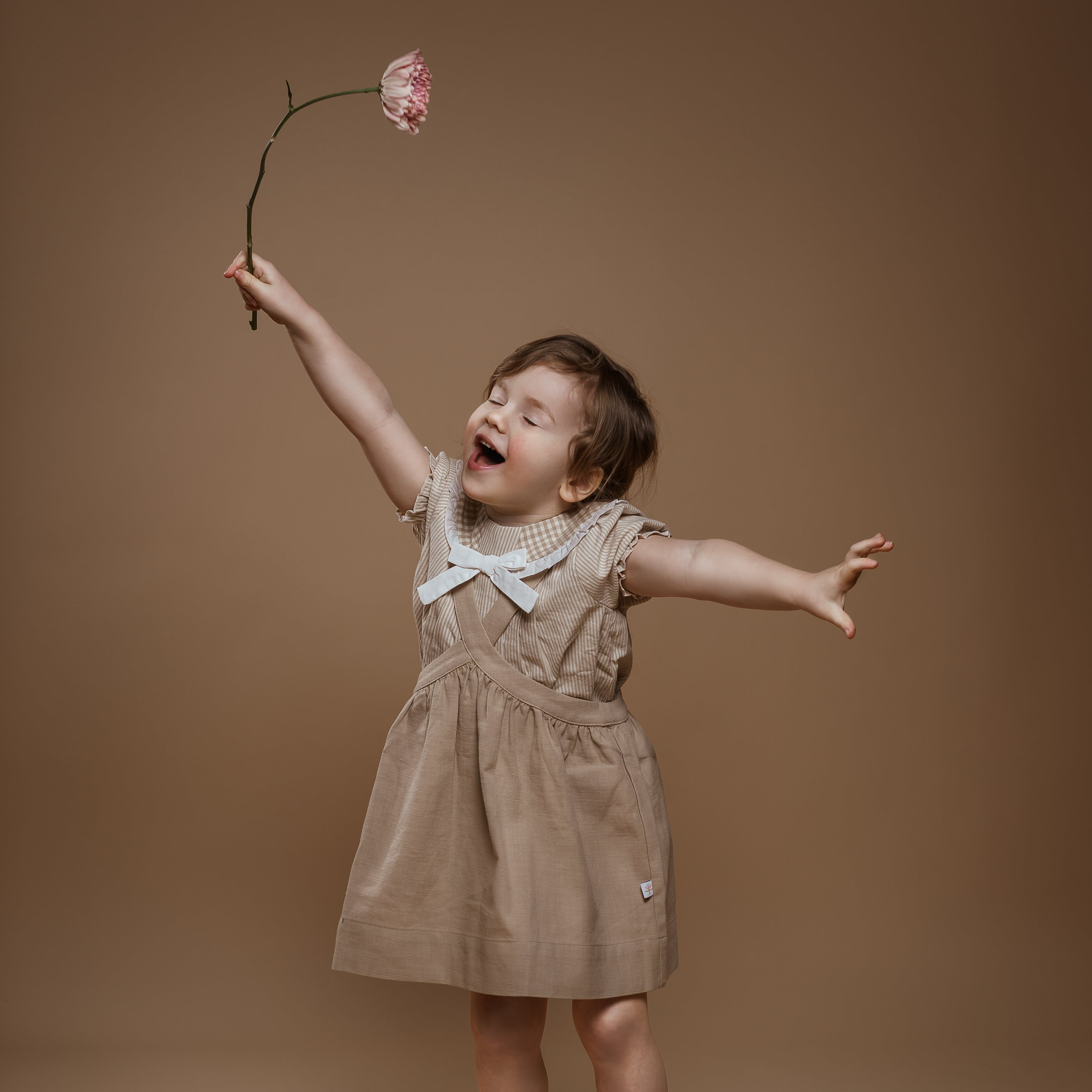 Child in a beige dress with red shoes holding a pink flower against a brown background