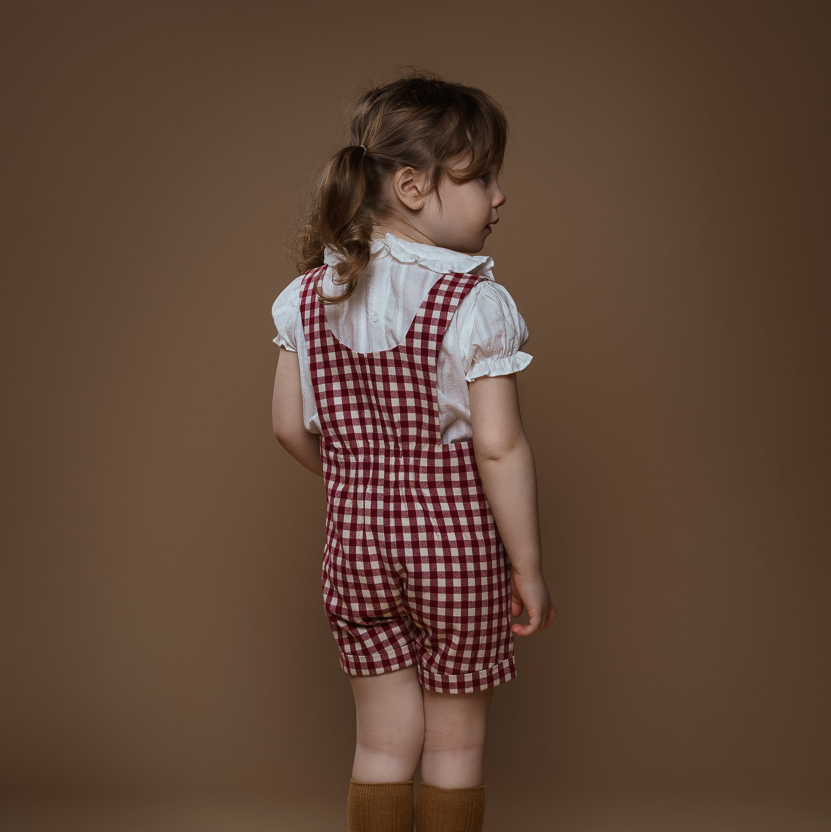 Child wearing a red checkered dress with a white shirt and brown boots on a brown background