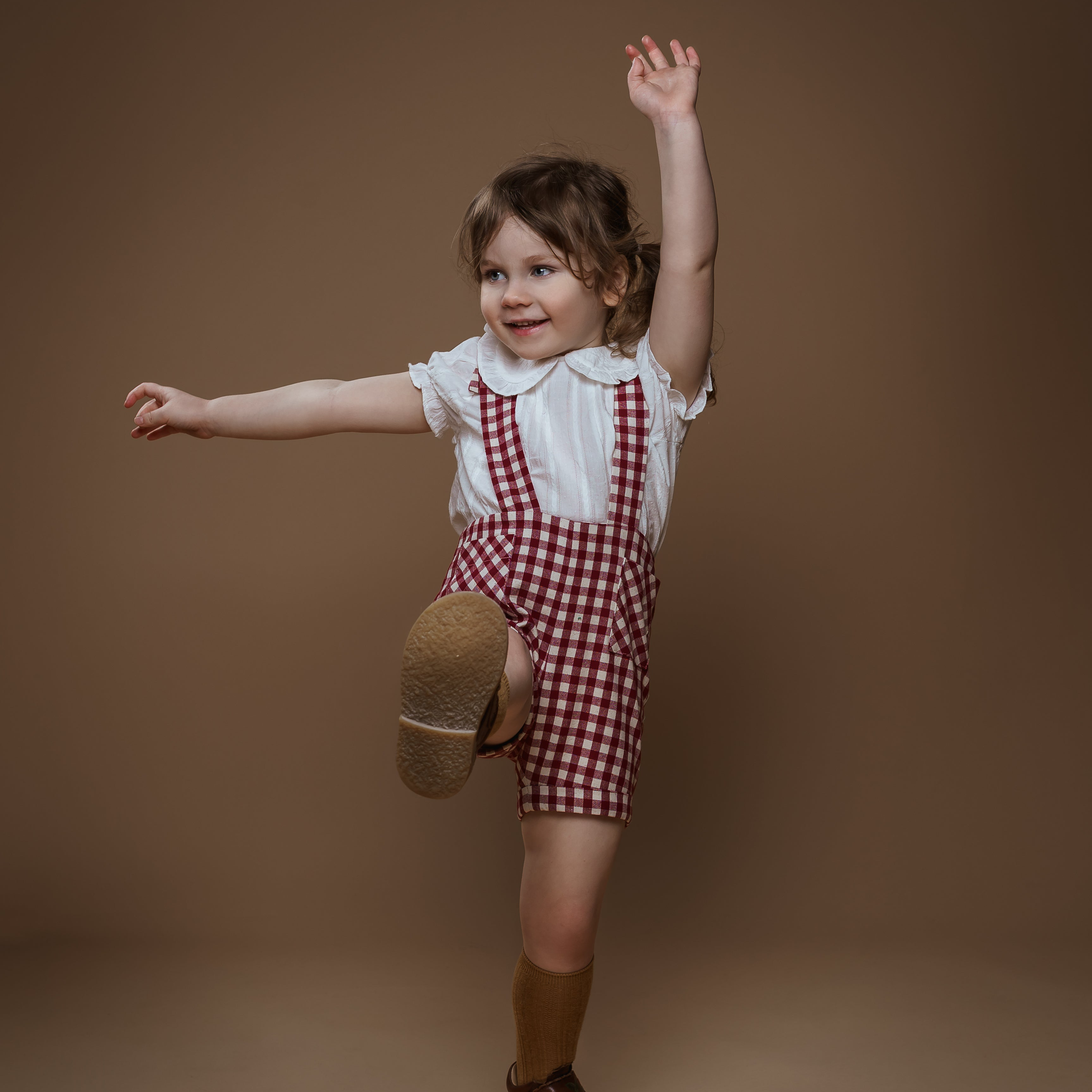 Child wearing a white shirt, red checkered overalls, and brown shoes on a brown background