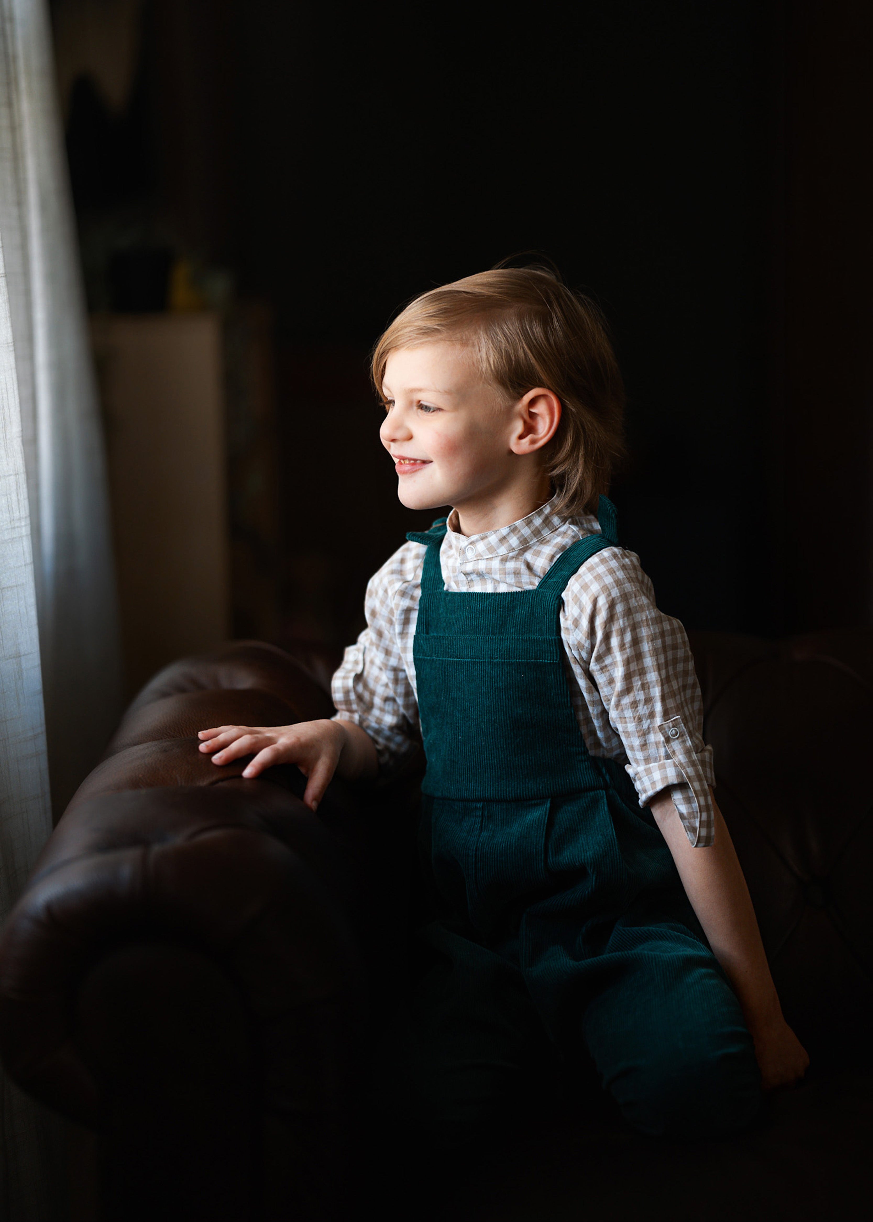 Young boy in a green dress sitting on a couch in a dimly lit room.