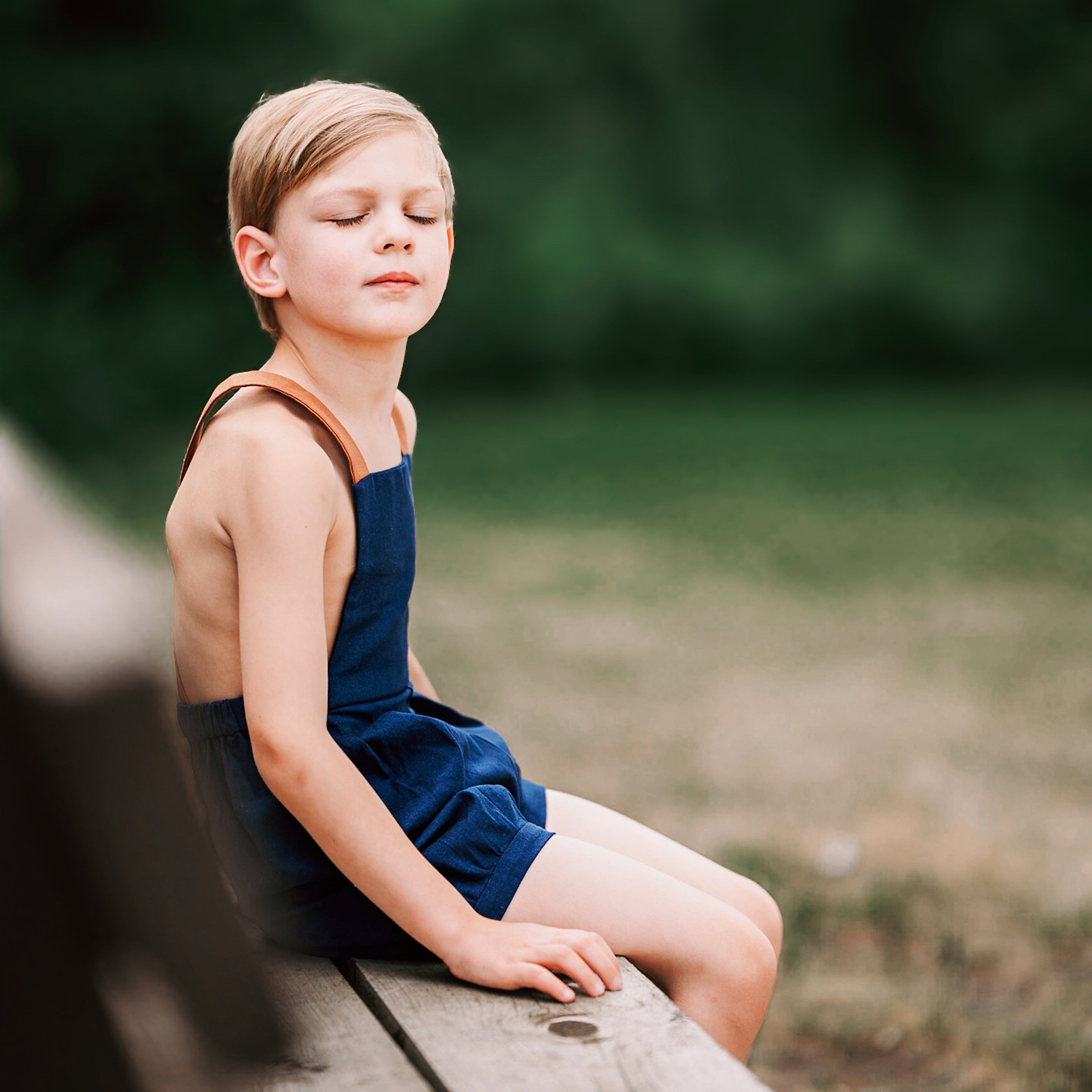 Boy with eye closed sitting on a bench wearing navy overalls