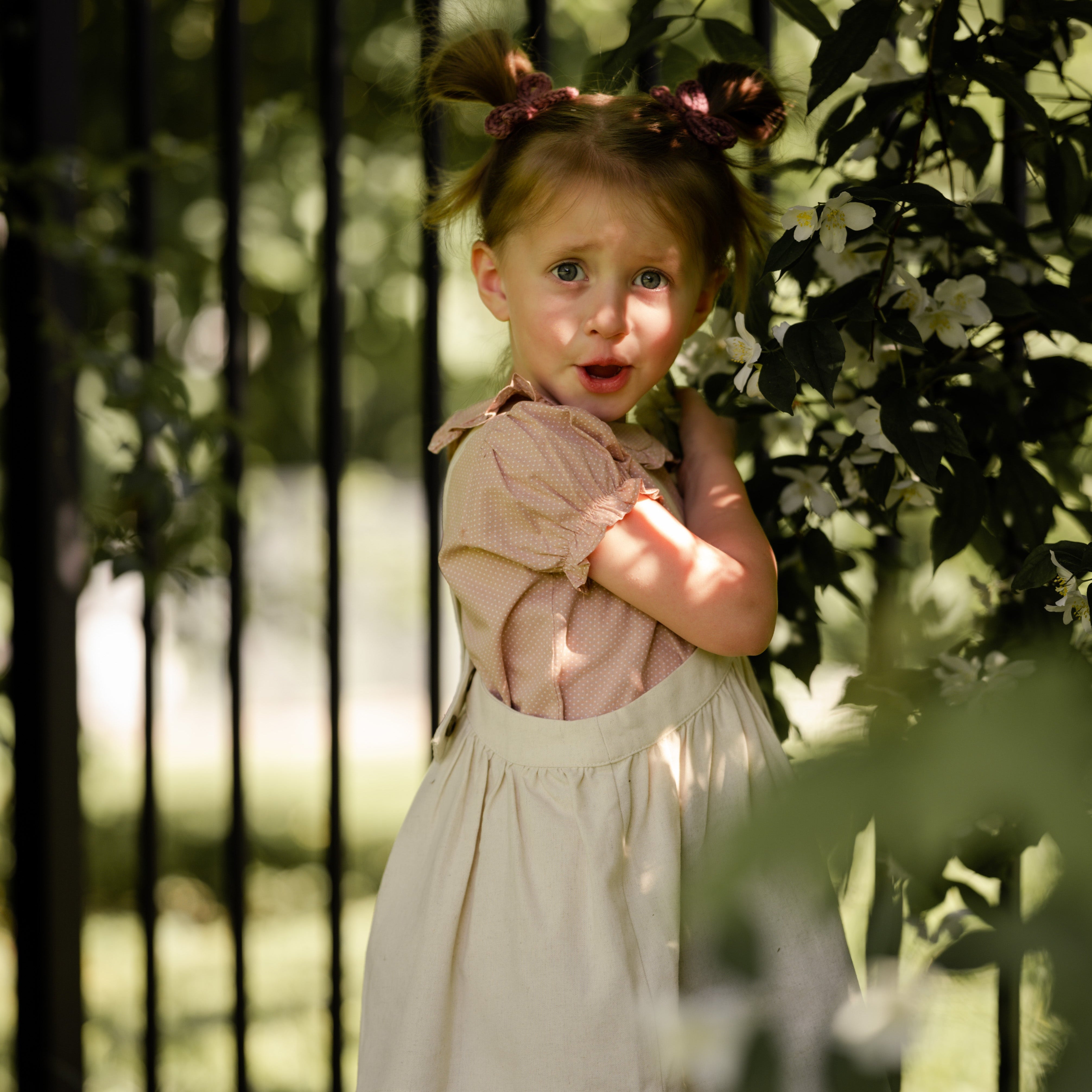 Young girl in a natural linen pinafore standing in front of a black metal gate with greenery around.