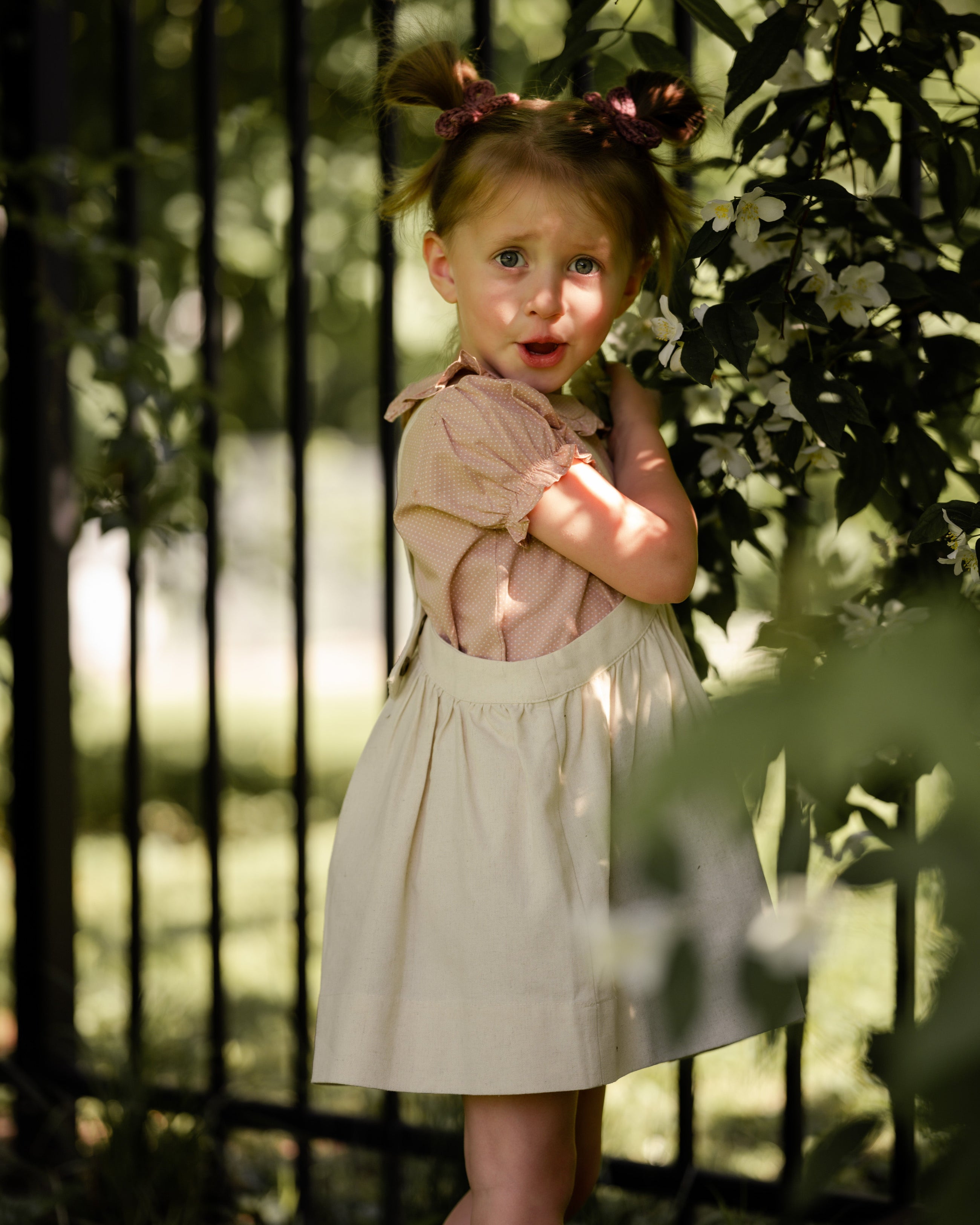 Young girl in a natural linen pinafore standing in front of a black metal gate with greenery around.