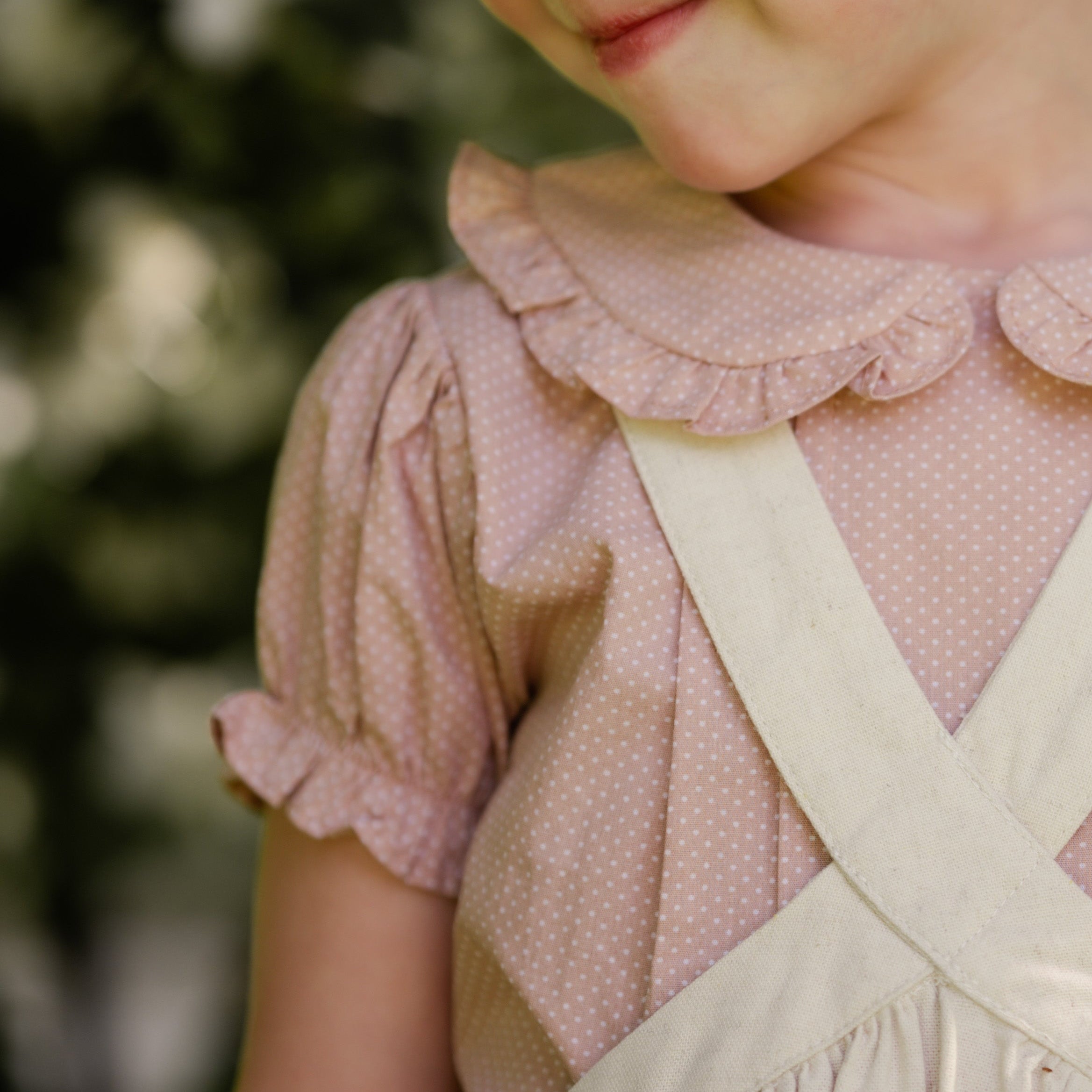 Close-up of a child wearing a beige blouse with ruffled collar and beige linen pinafore.