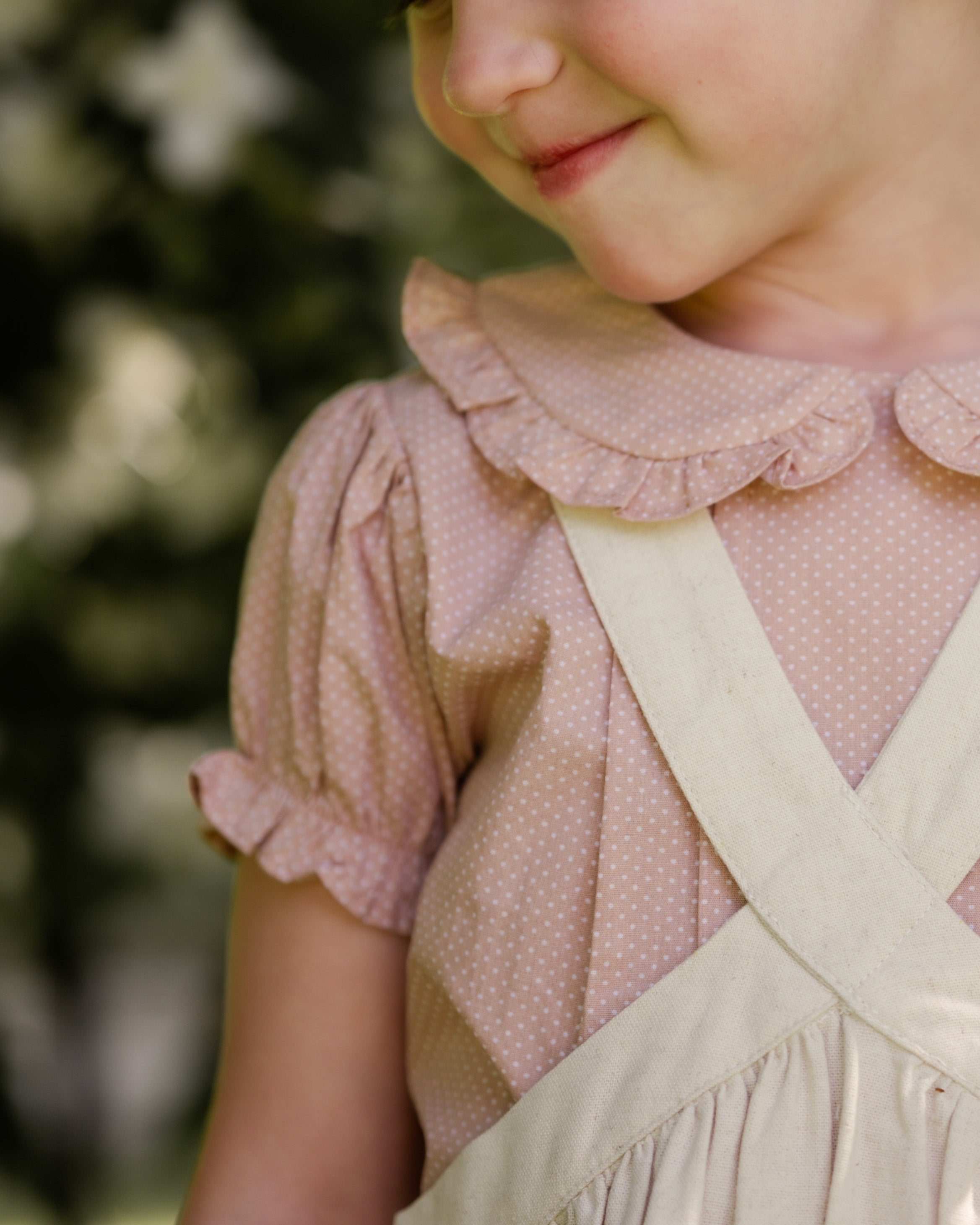 Close-up of a child wearing a beige blouse with ruffled collar and beige linen pinafore.