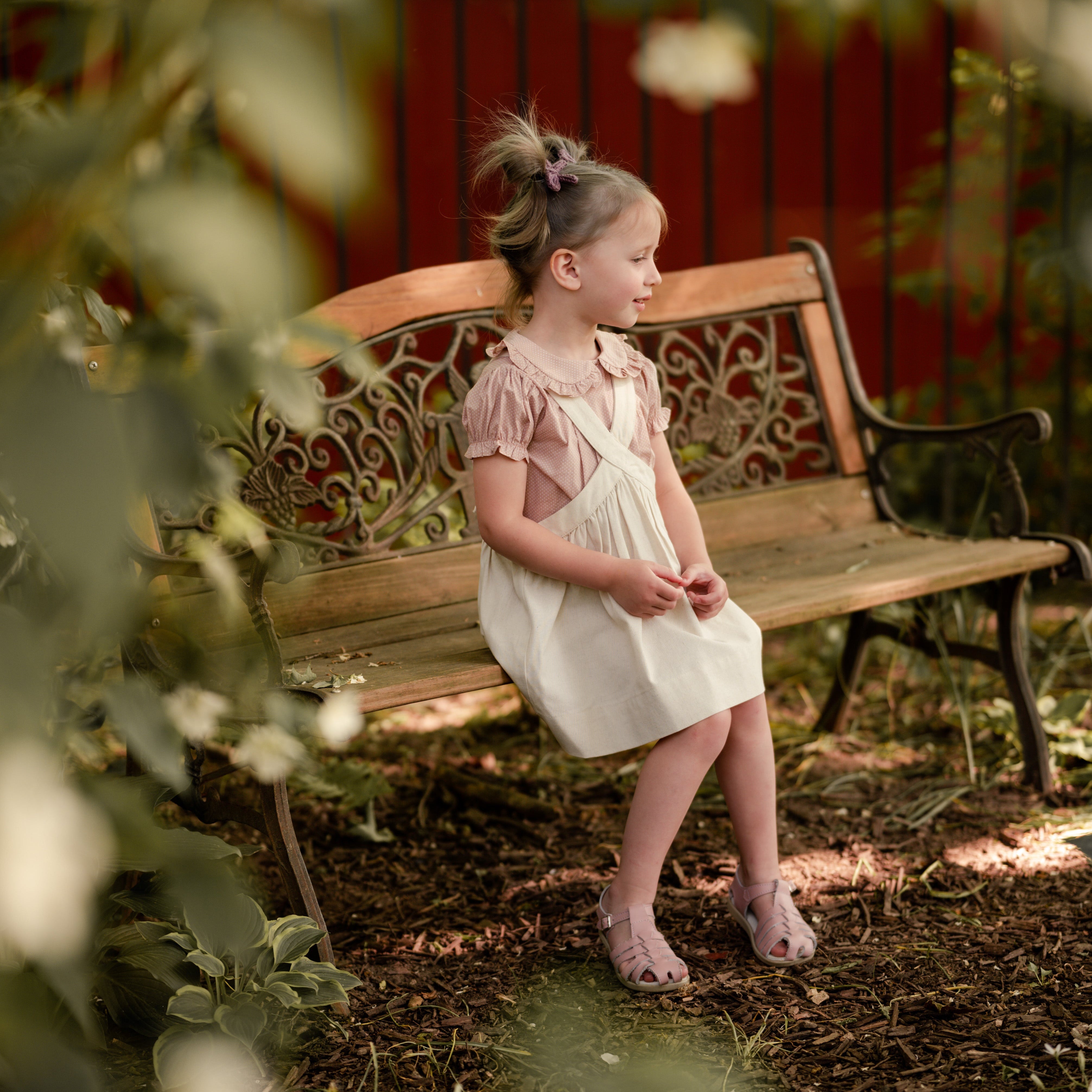 Young girl in a natural linen pinafore dress sitting on a wooden bench in a garden.