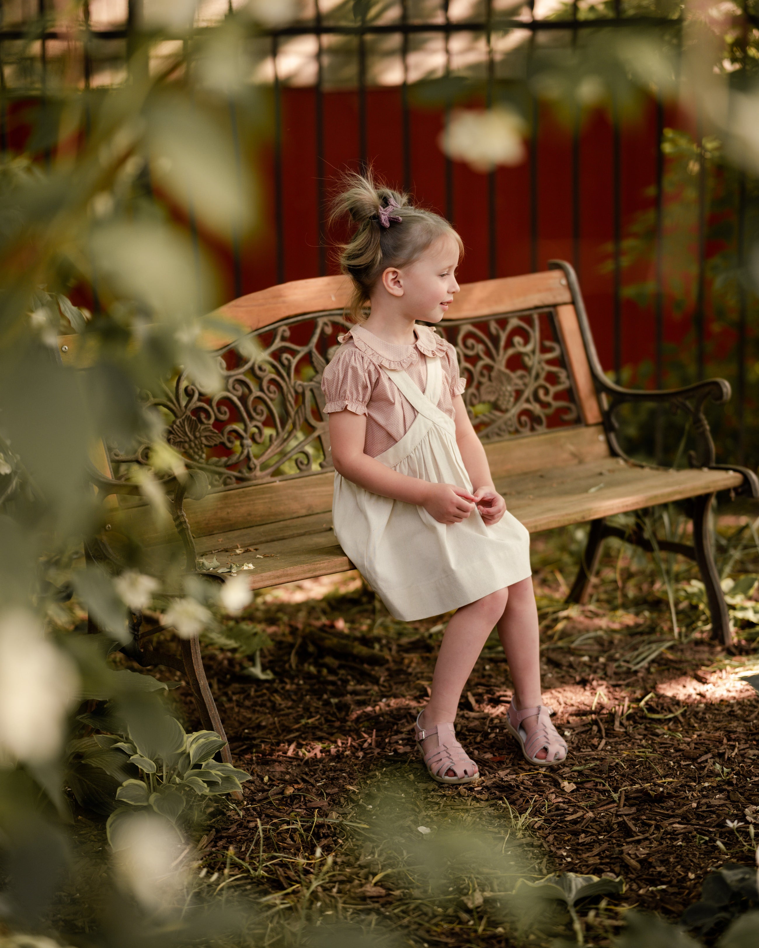 Young girl in a natural linen pinafore dress sitting on a wooden bench in a garden.