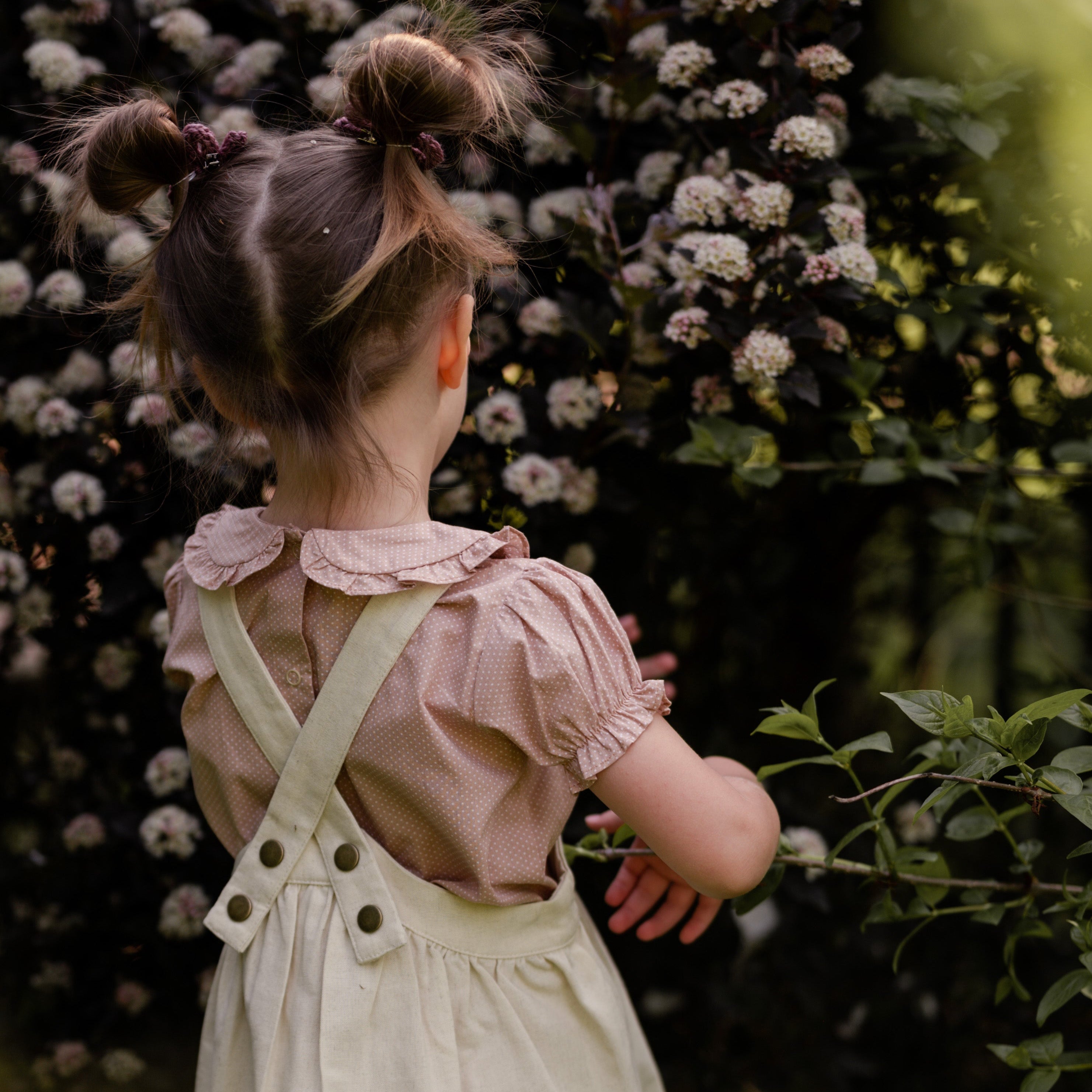 Girl wearing a beige blouse and beige pinafore with suspenders standing in front of a floral background