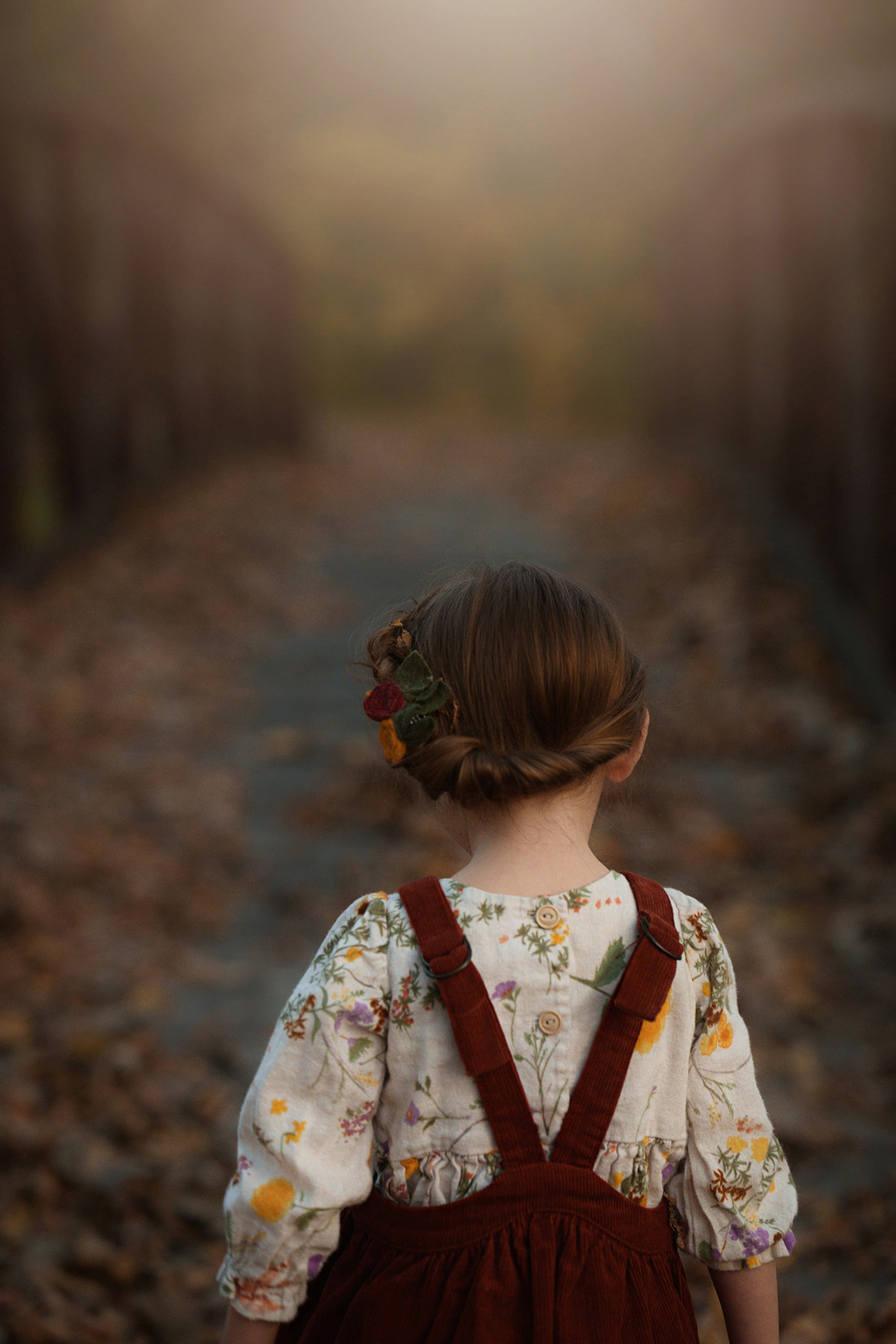 Child in a floral blouse and brown overalls standing in a forest with a blurred background