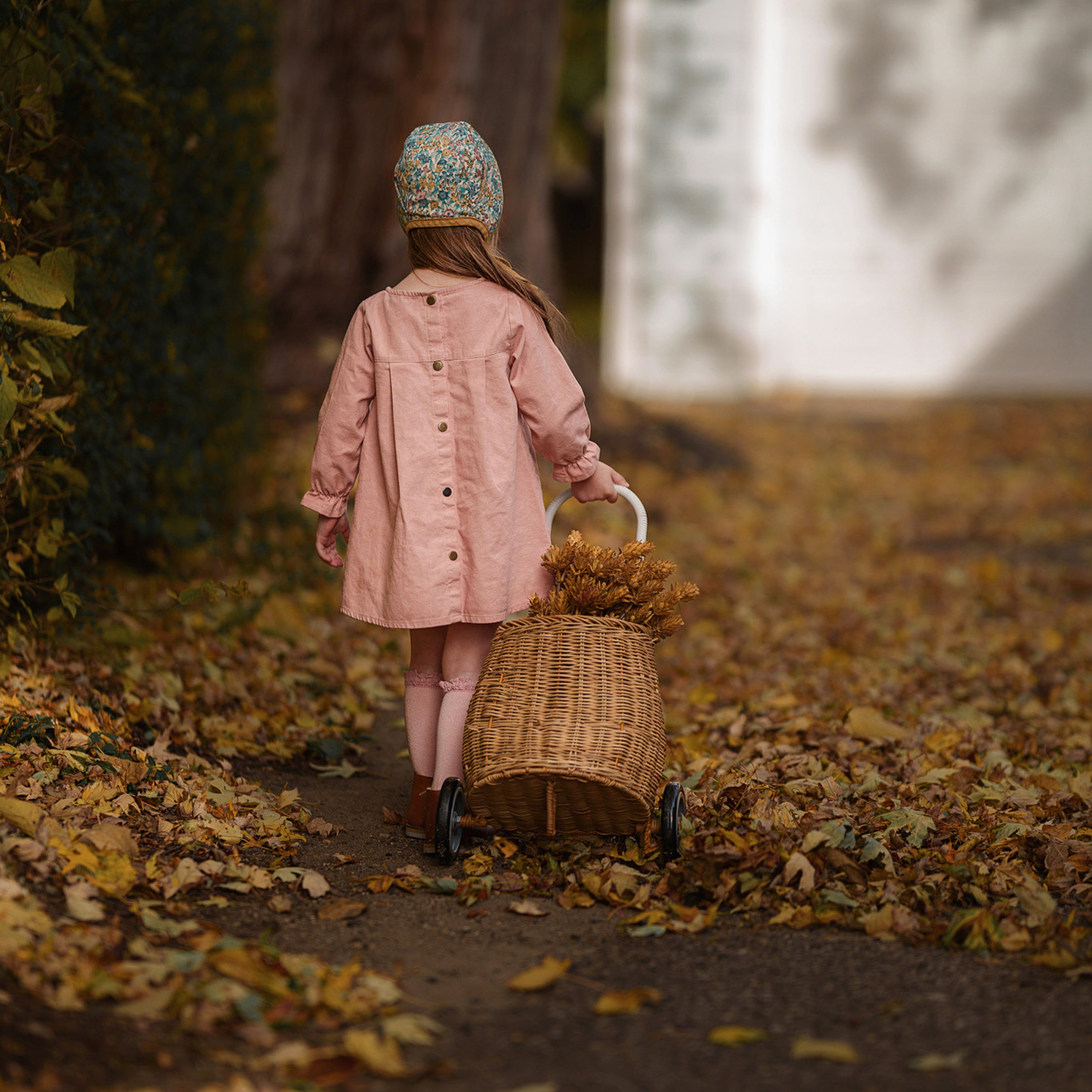 Child in a pink coral dress with long sleeves walking with a wicker basket on a leaf-covered path.