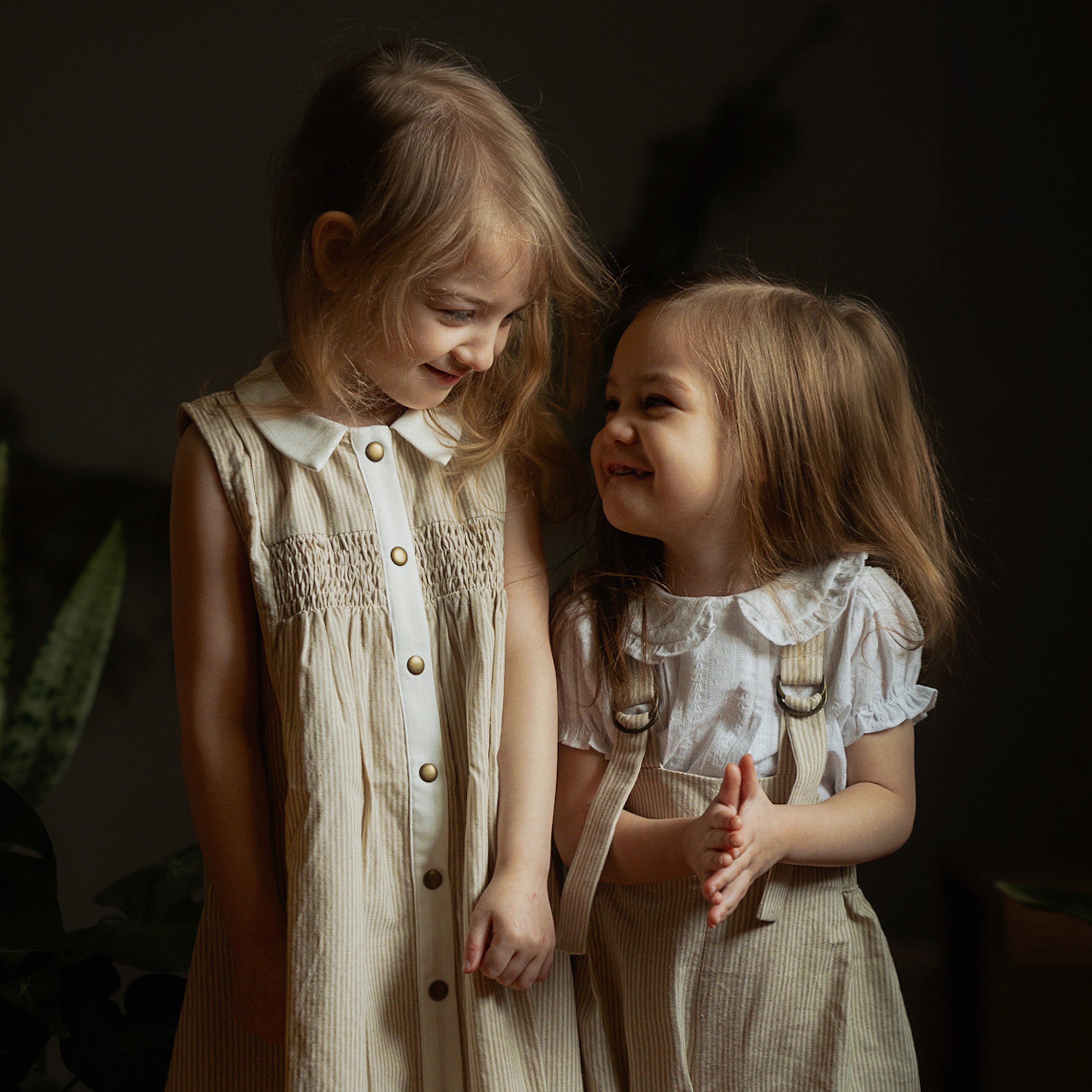 Two little girls wearing beige stripes Grow-With-Me Rompers and a matching beige stripes dress, playing together indoors, highlighting the adjustable straps, soft fabric, and coordinated timeless designs.