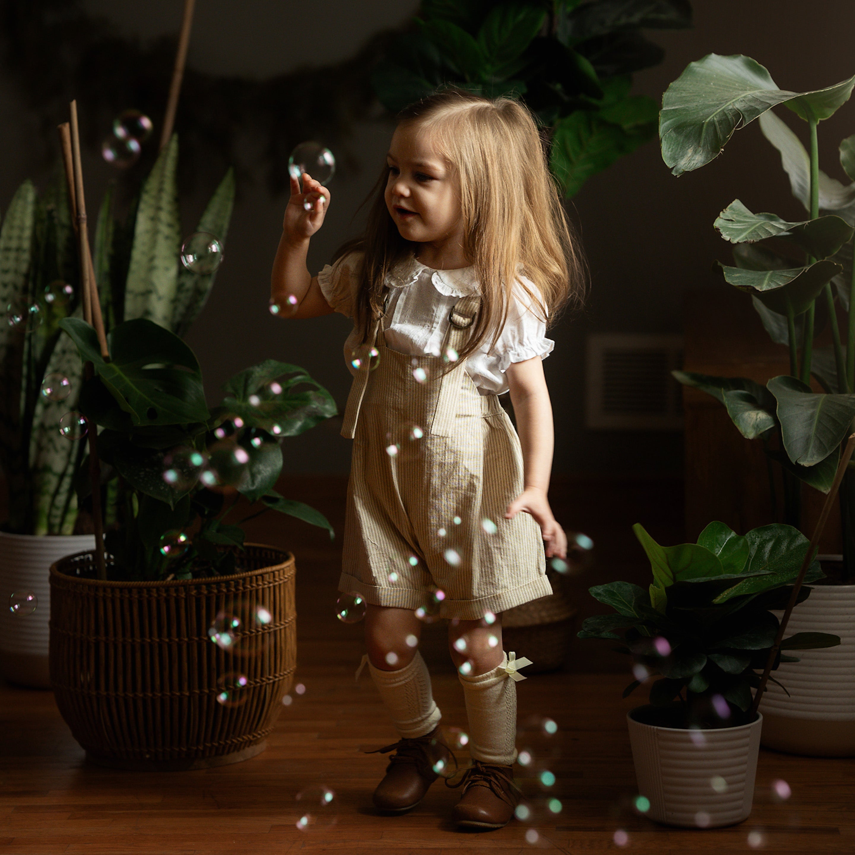 A tiny 2-year-old wearing a beige stripes Grow-With-Me Romper over a frilly white heirloom style blouse, playing indoors with soap bubbles in a cozy setting, showcasing the adjustable straps and soft fabric of the romper paired with the elegant details of the blouse.