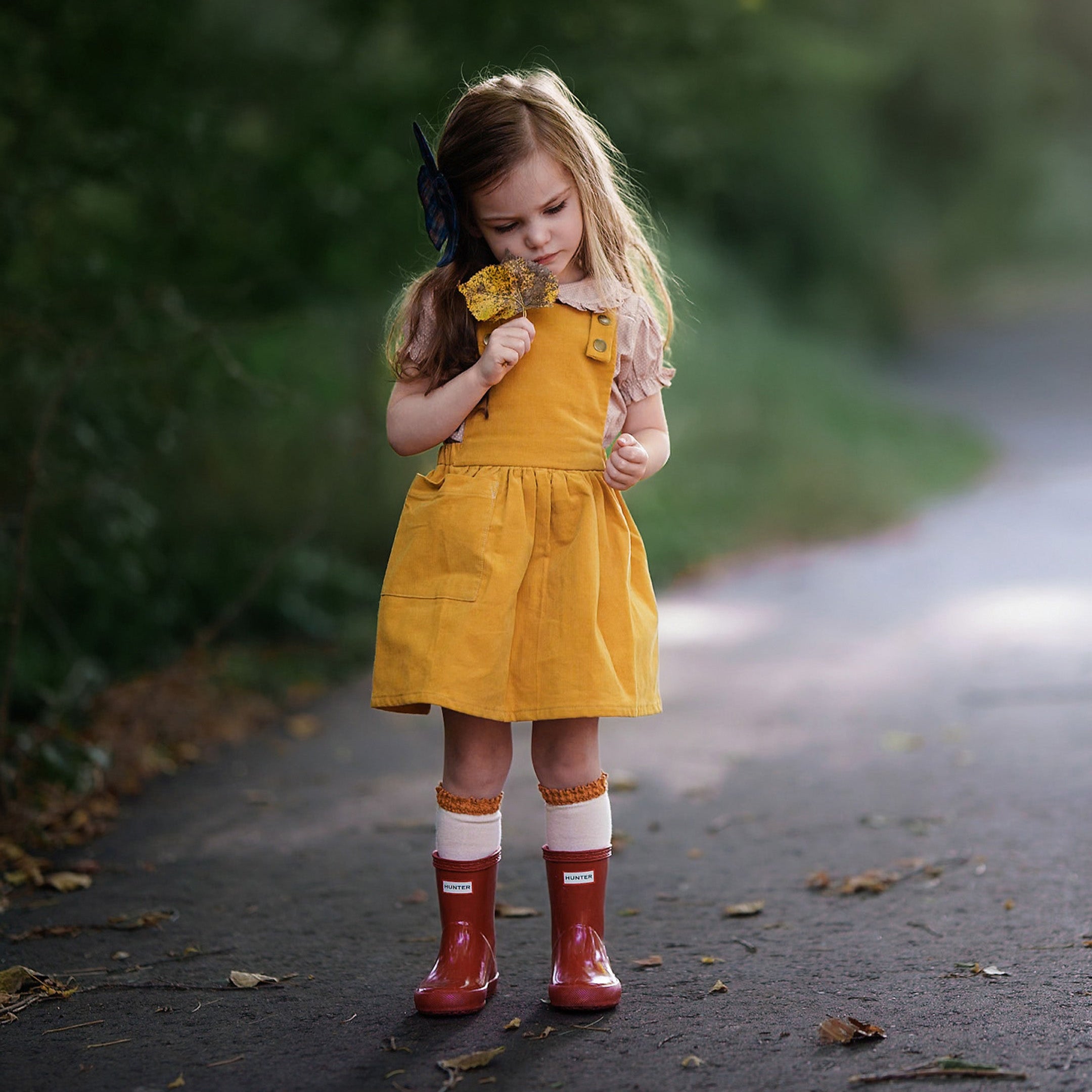 Model girl standing wearing rain boots and 3-year-old girl modeling the Mustard Pinafore paired with rain boots and a frilly beige dots blouse, highlighting a playful summer look that combines style and comfort.