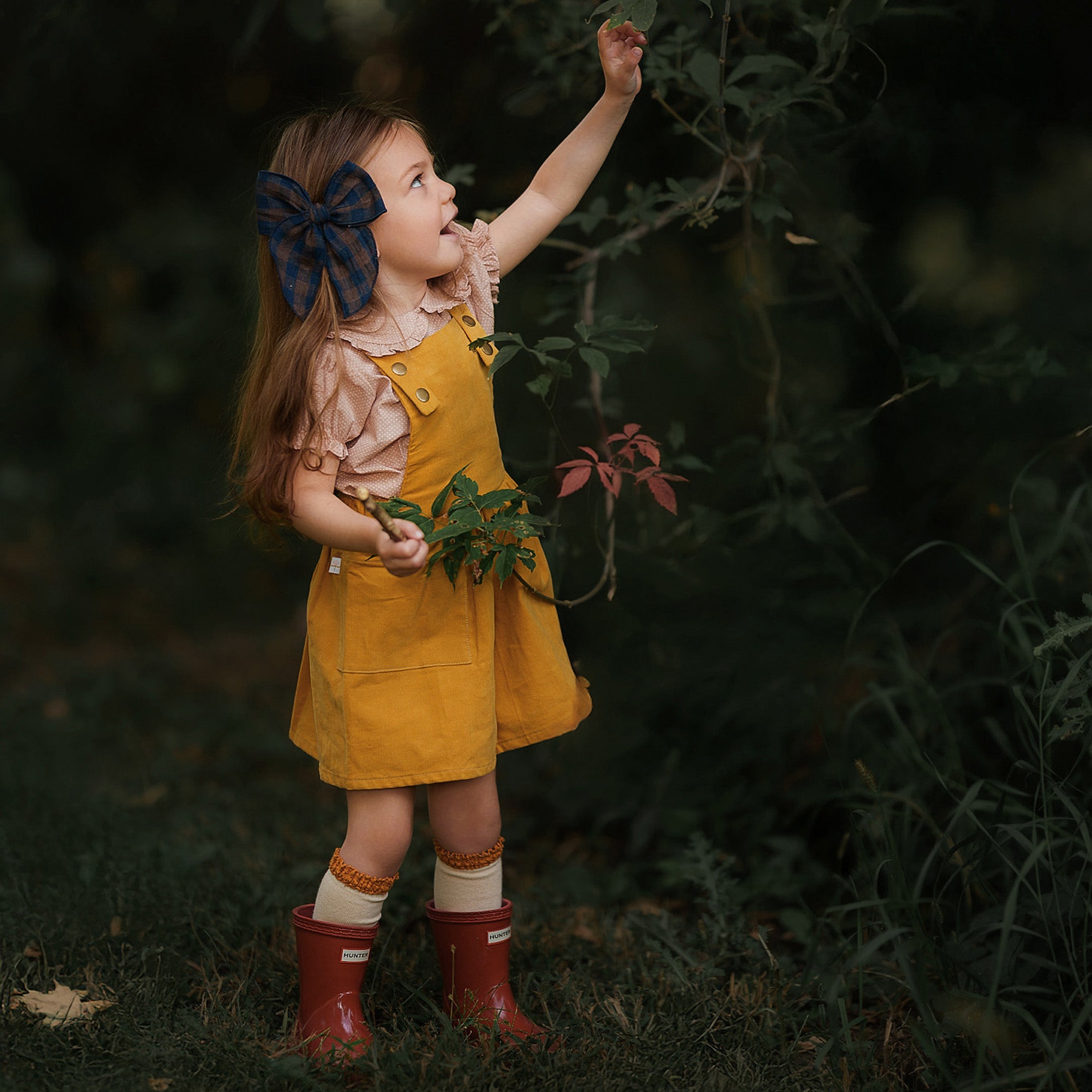 Side view of a girl in the Mustard Pinafore, joyfully picking flowers, demonstrating the garment’s versatility and how it can be worn on its own during summer adventures.