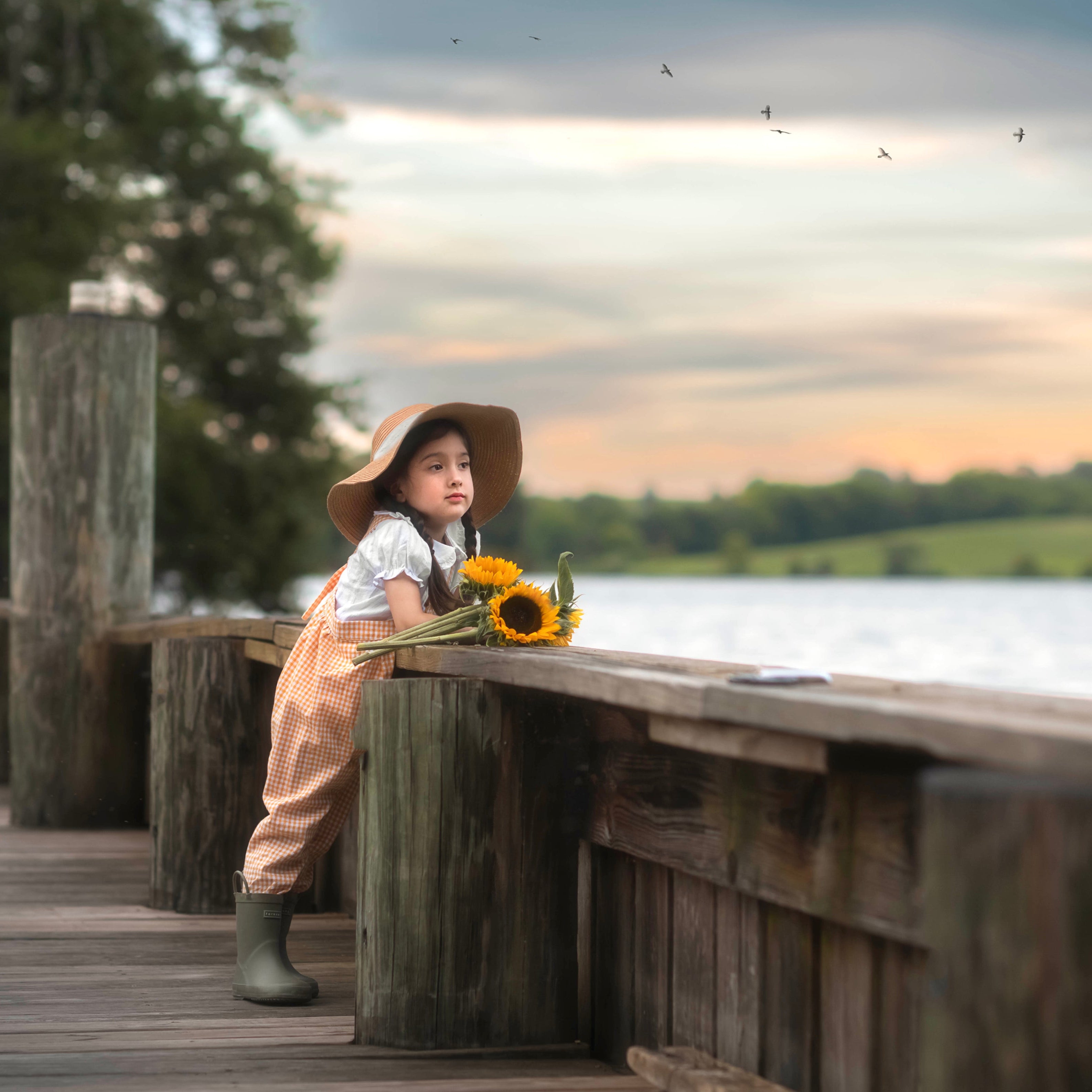 Girl on a wooden dock by a lake with sunflowers, wearing a hat and rain boots.