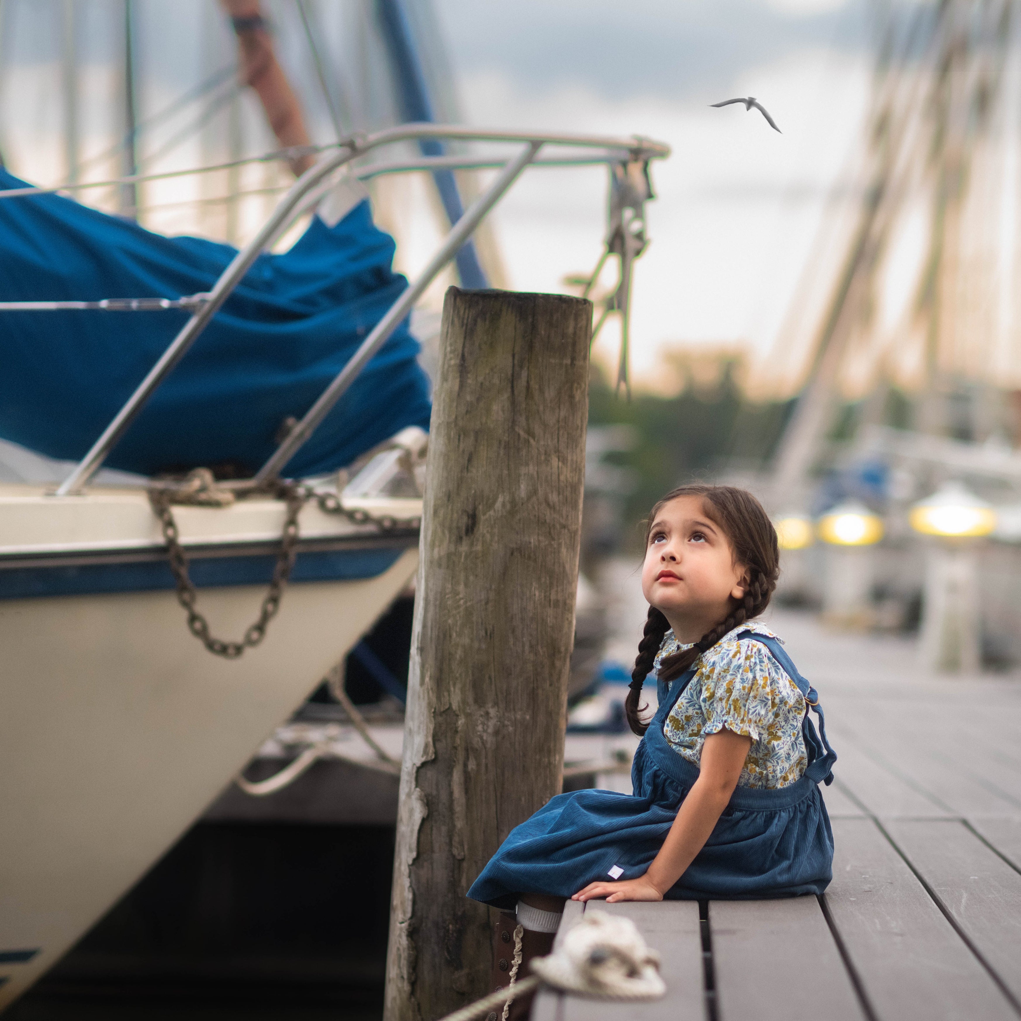 Young girl sitting on a dock by a boat, looking up at the sky.