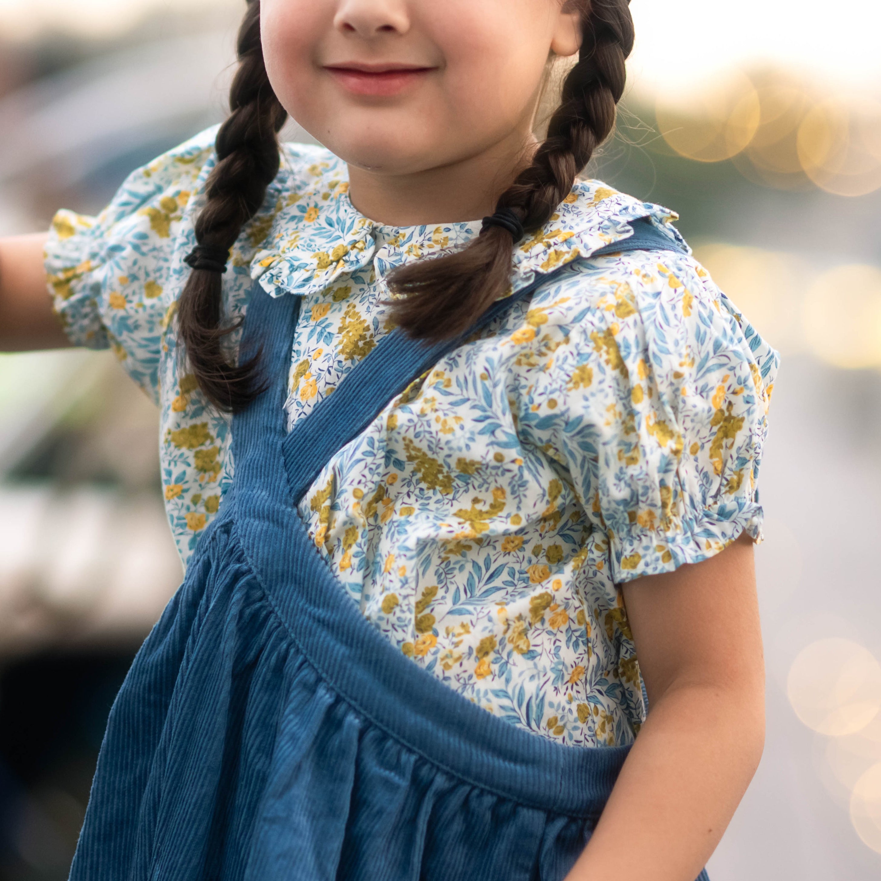 Young girl in a floral blouse and blue overalls standing outdoors with blurred background