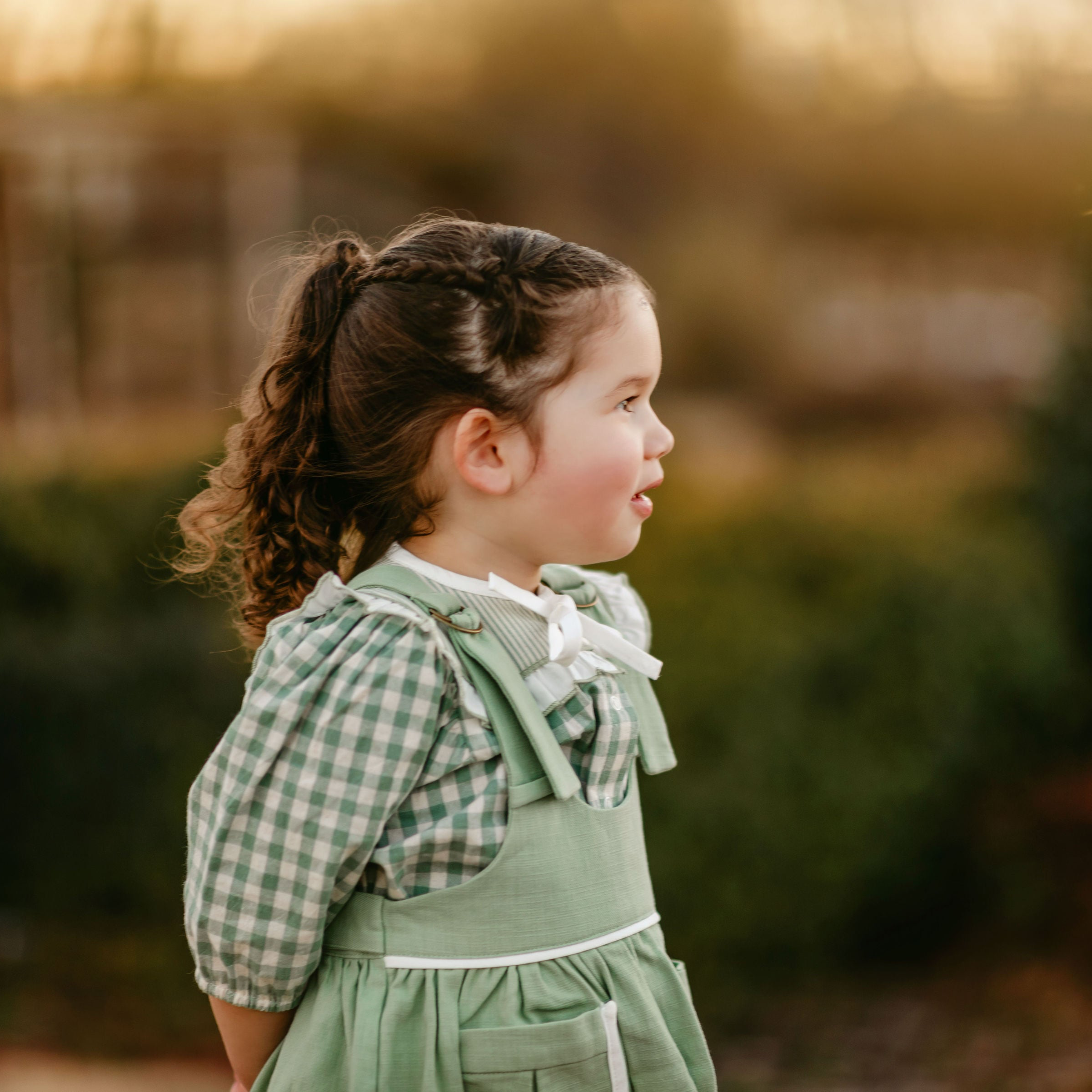 Young girl in a green checkered dress standing outdoors with a blurred background