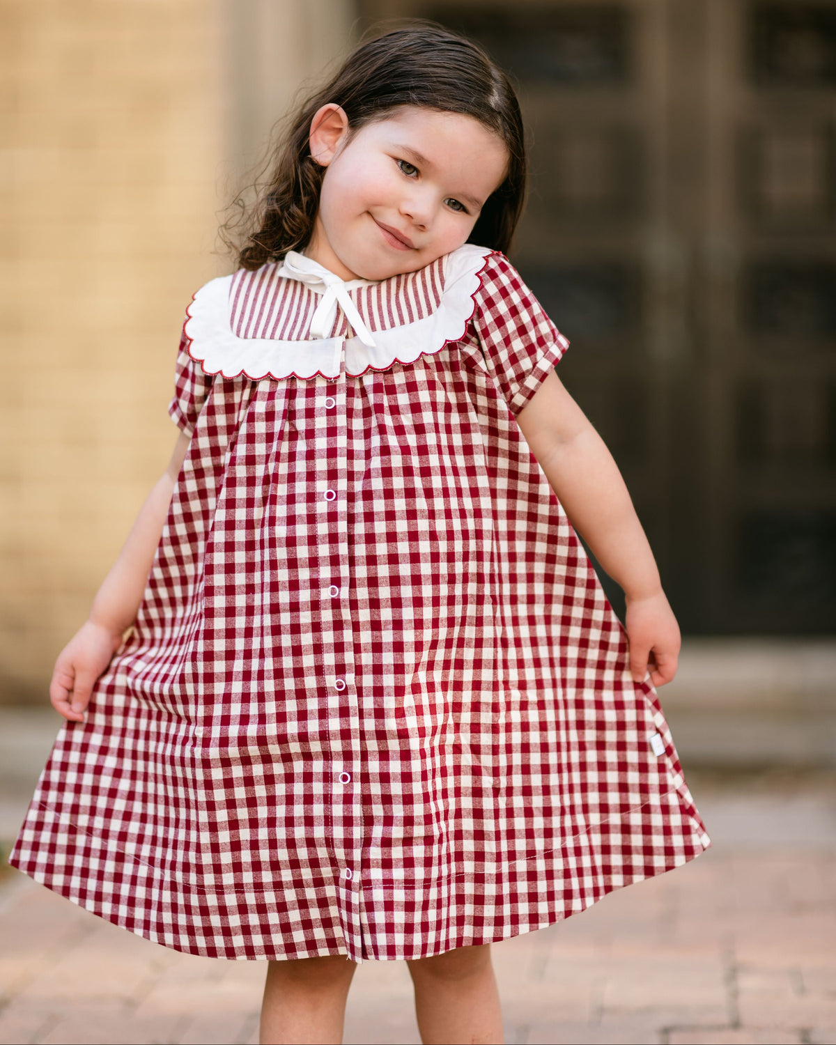 Young girl wearing a red and white checkered dress with a white collar, standing outdoors.