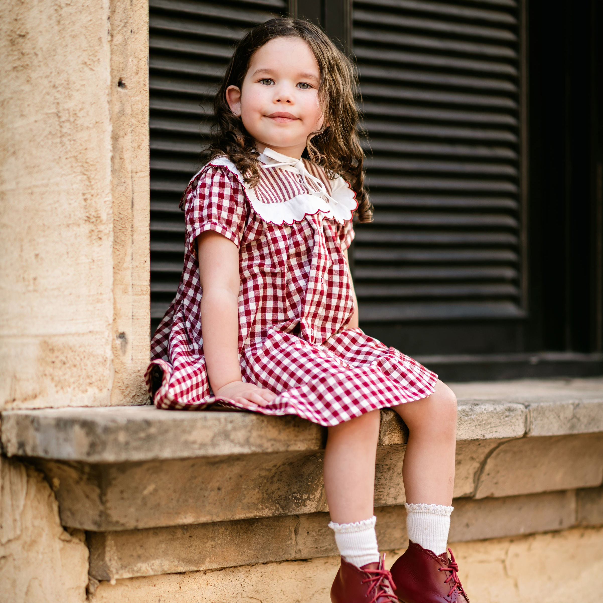 Young girl in a red checkered dress sitting on a stone ledge.
