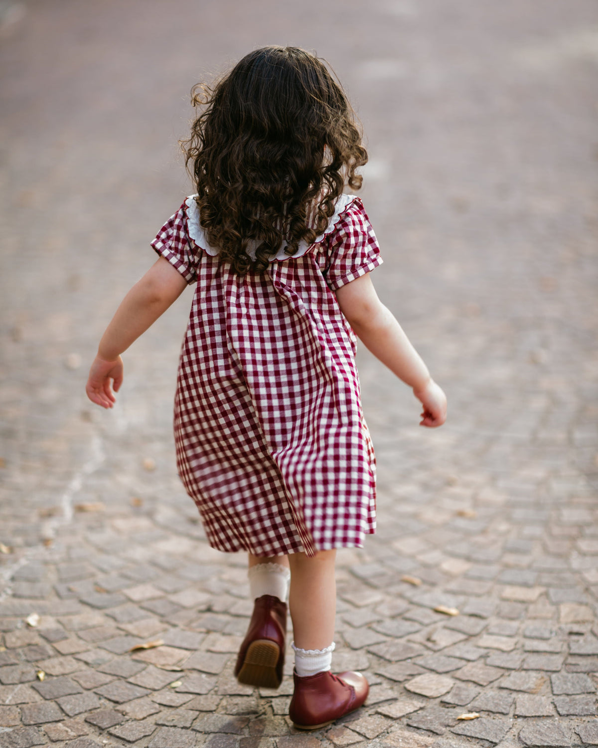 Child in a red and white checkered dress walking on a cobblestone street.