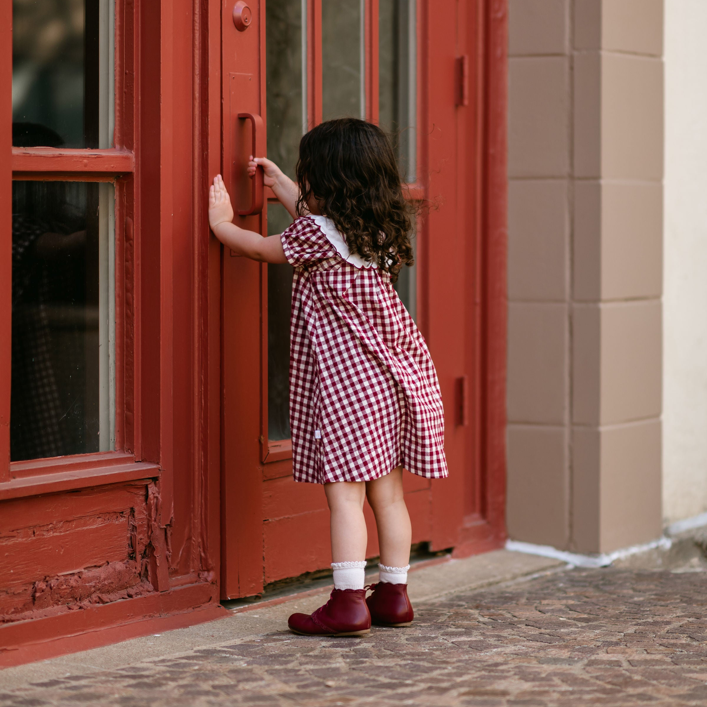 Child in a red and white checkered dress standing in front of a red door.