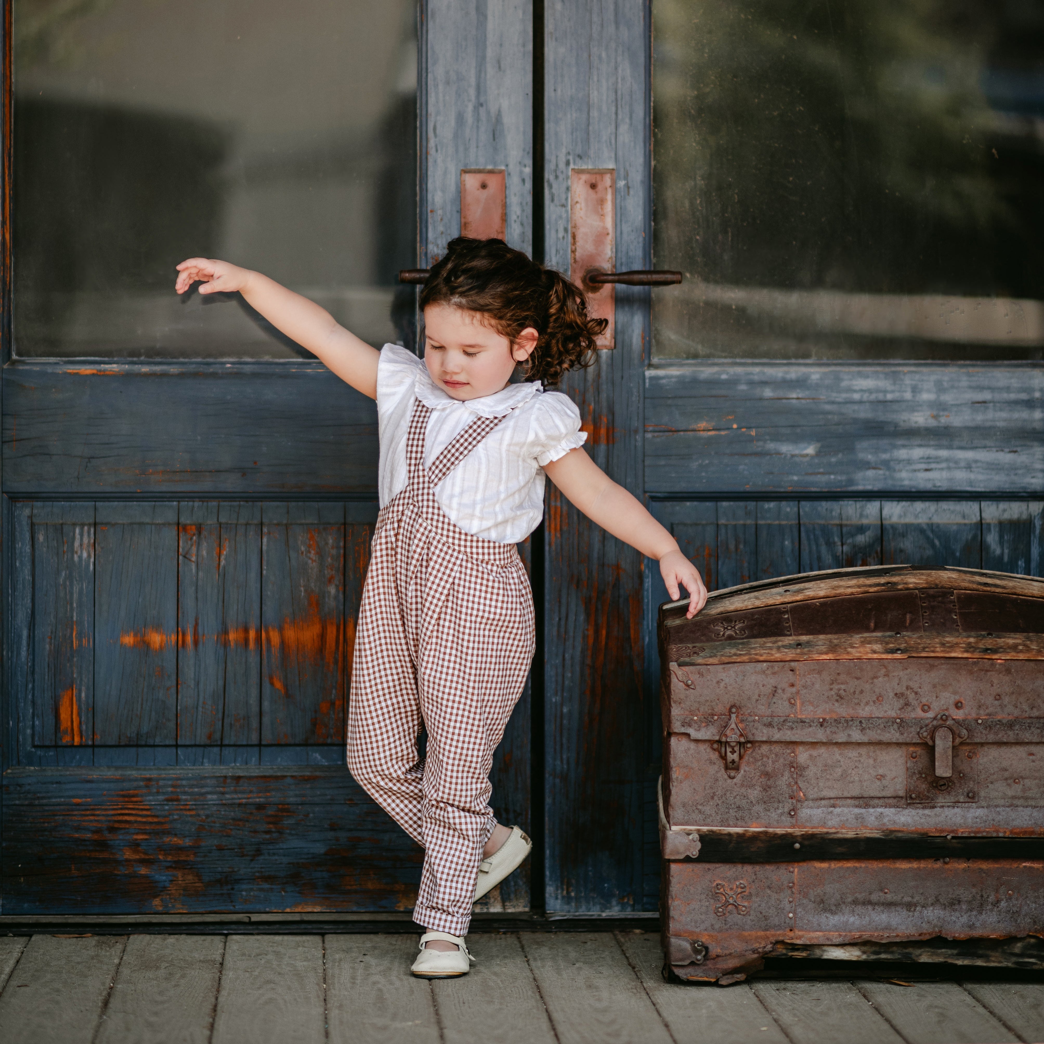 Child in a brown checkered jumpsuit standing in front of a large door with 'Daily News' written on it.