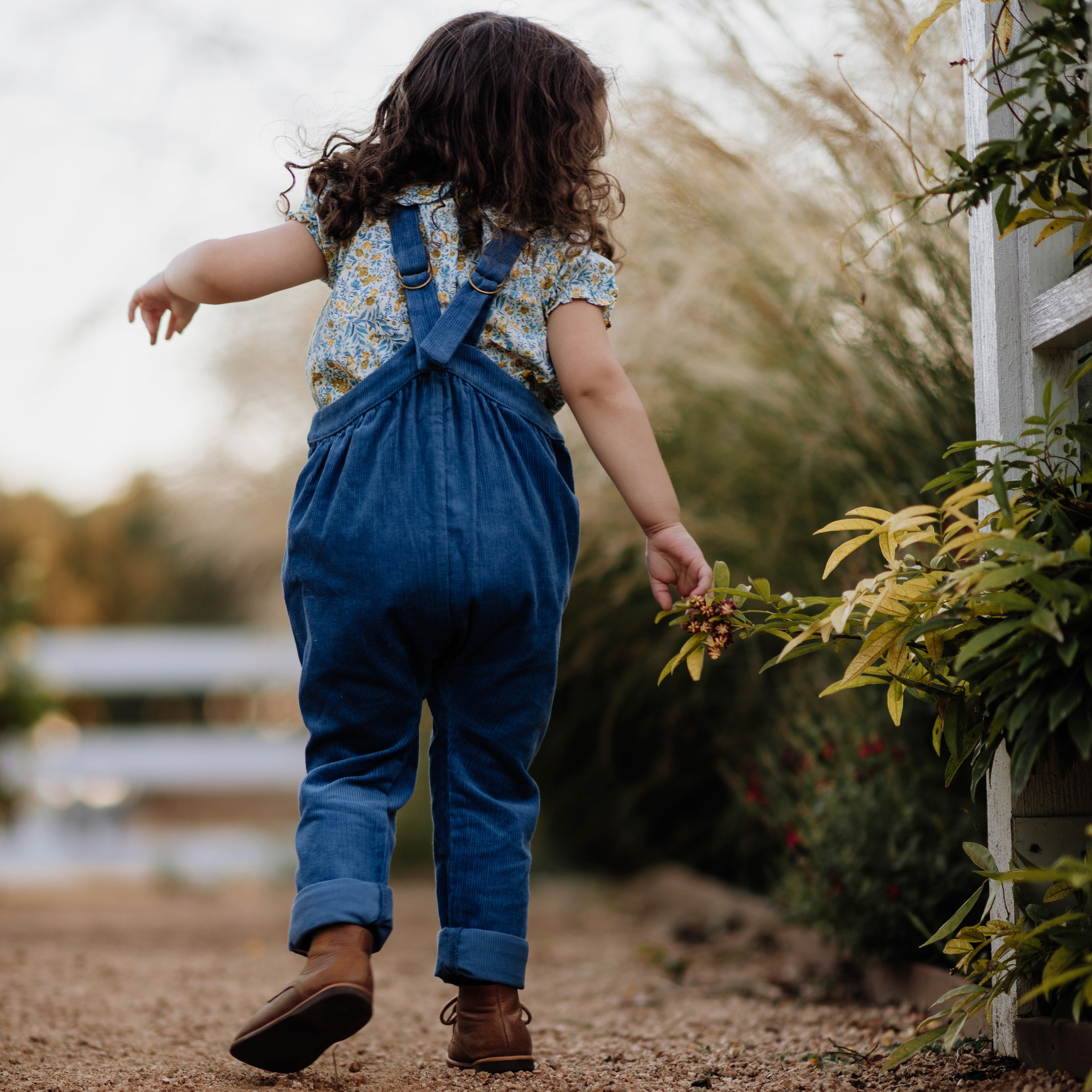 Child in blue overalls walking on a dirt path with greenery around