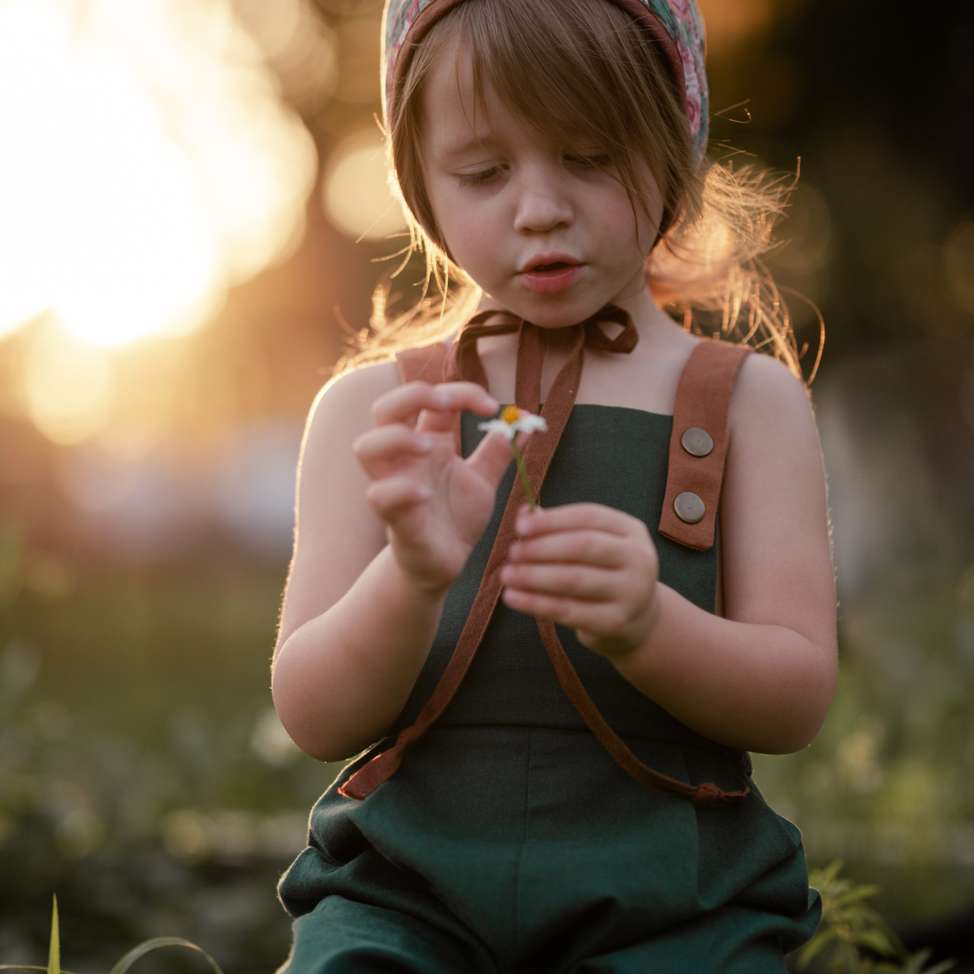 A 3-year-old girl sitting down, wearing the evergreen long overalls and a cute bonnet, playfully holding a daisy, highlighting comfort and charm in children’s fashion.