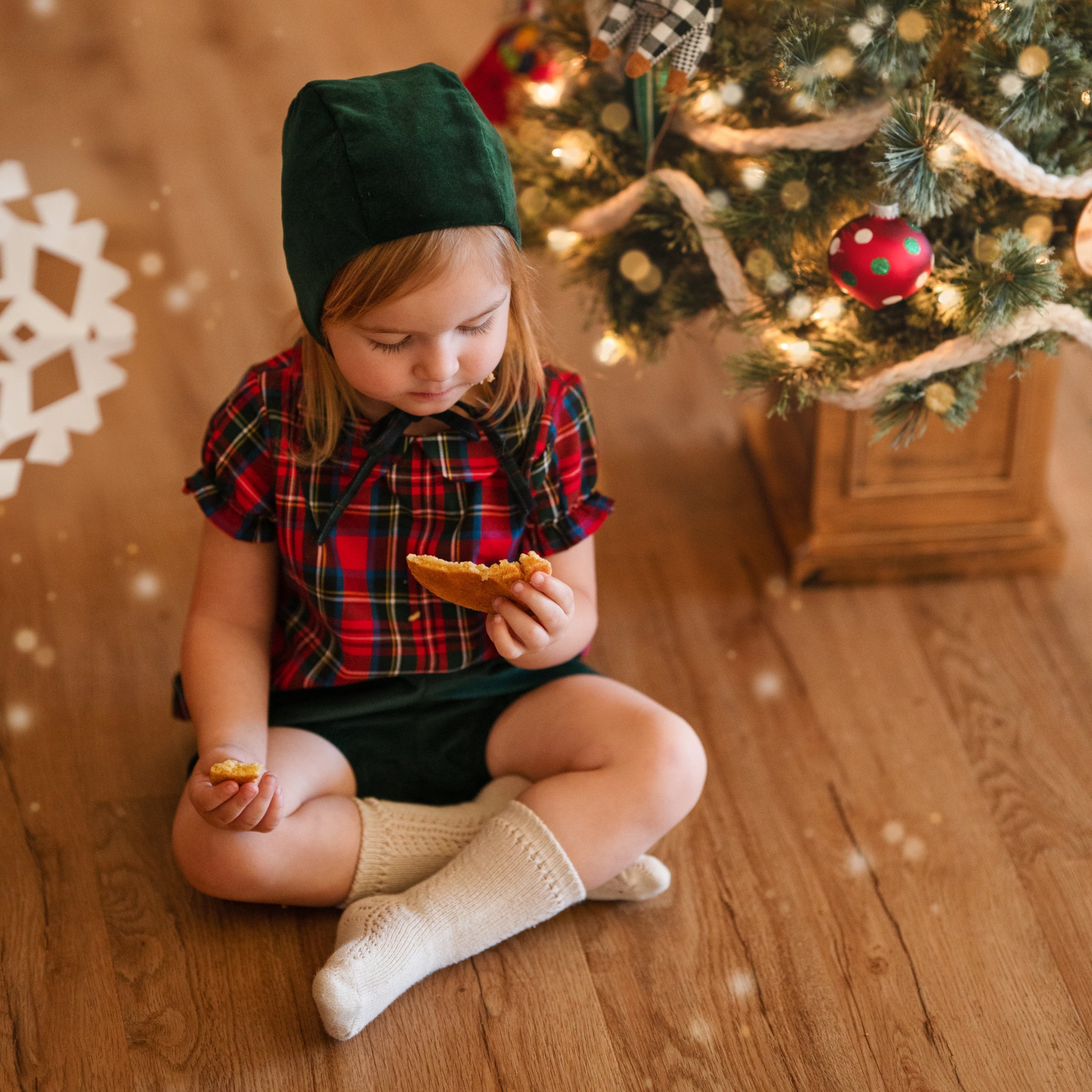 Girl in green bonnet and plaid blouse eating cooking in front of a Xmas tree