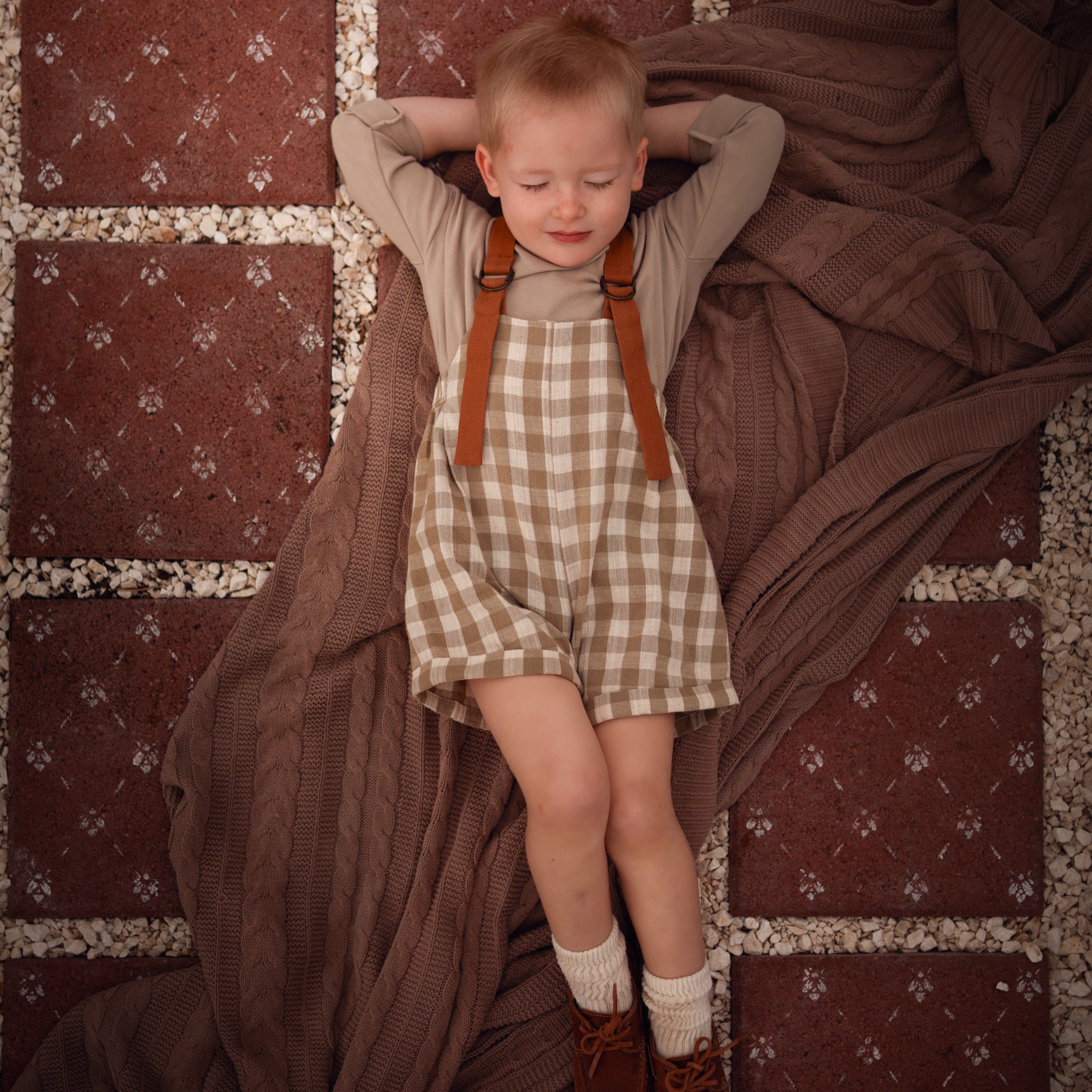 Model boy lying down in a Grow-With-Me Romper in Beige Gingham over a neutral top, showcasing the adjustable straps and classic gingham pattern, exuding a relaxed and playful vibe while emphasizing the romper's versatility and comfort.