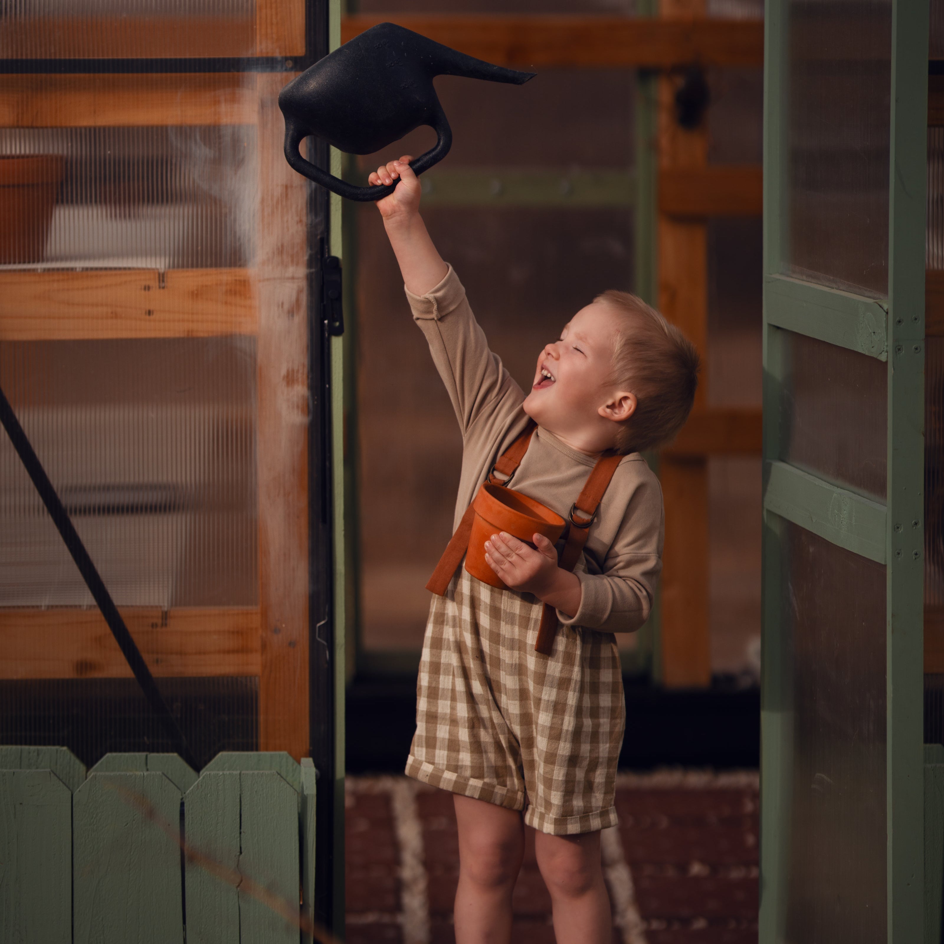 Model boy playing with a watering can in front of a green door, wearing the Grow-With-Me Romper in Beige Gingham over a neutral top, laughing joyfully as he enjoys a playful moment, highlighting the romper's comfort and style in a cheerful outdoor setting.