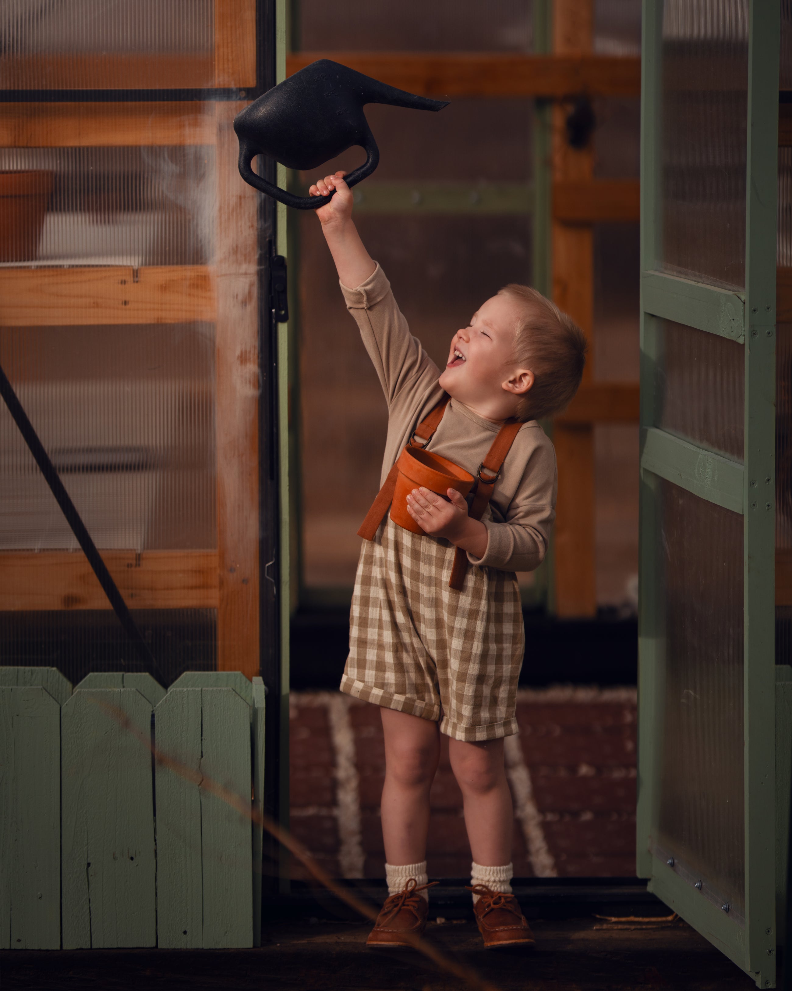 Model boy playing with a watering can in front of a green door, wearing the Grow-With-Me Romper in Beige Gingham over a neutral top, laughing joyfully as he enjoys a playful moment, highlighting the romper's comfort and style in a cheerful outdoor setting.