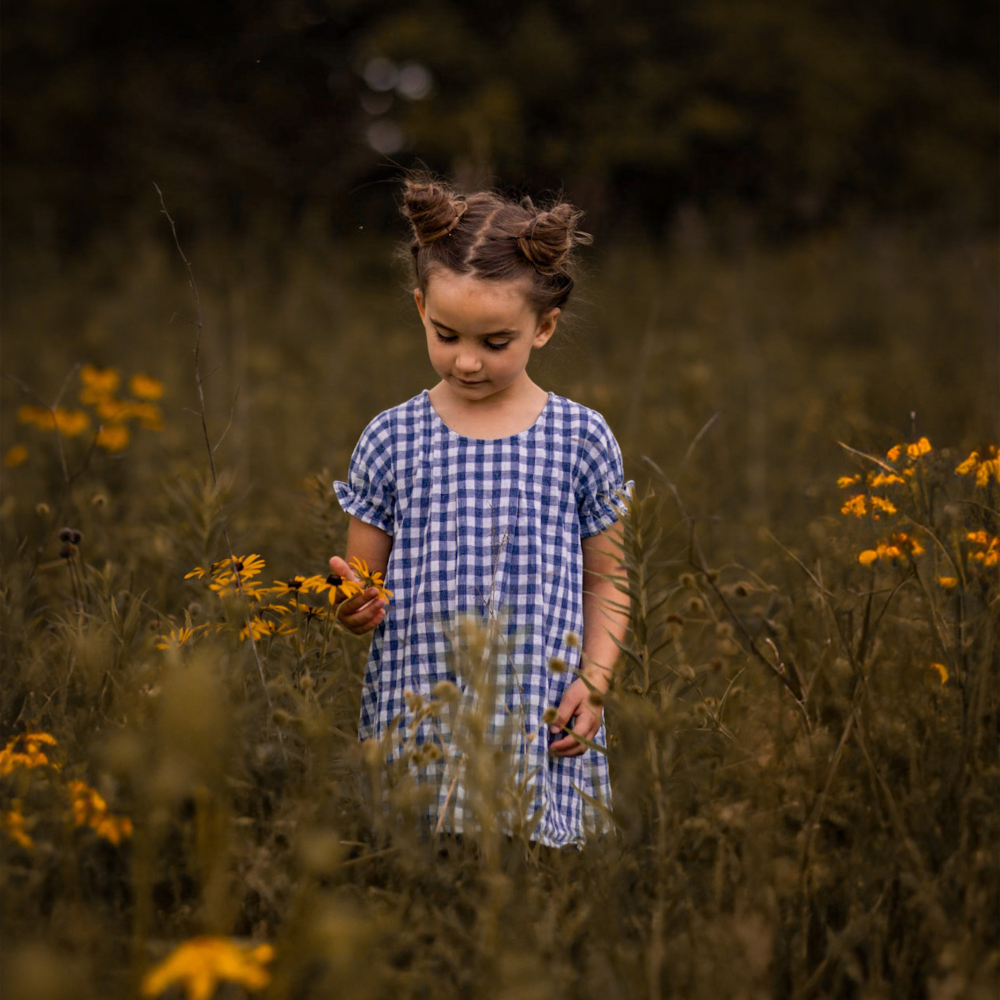 A little girl stands among vibrant flowers, wearing a Blue Gingham linen dress. The back view of the dress reveals its pin-tucked design, a yoke with pleats, and delicate ruffles at the short sleeve hem. Surrounded by colorful blooms, she embodies a joyful spirit, with her hair styled up, enjoying the beauty of nature. The A-line shape of the dress allows for comfort and ease as she plays in this enchanting floral setting.