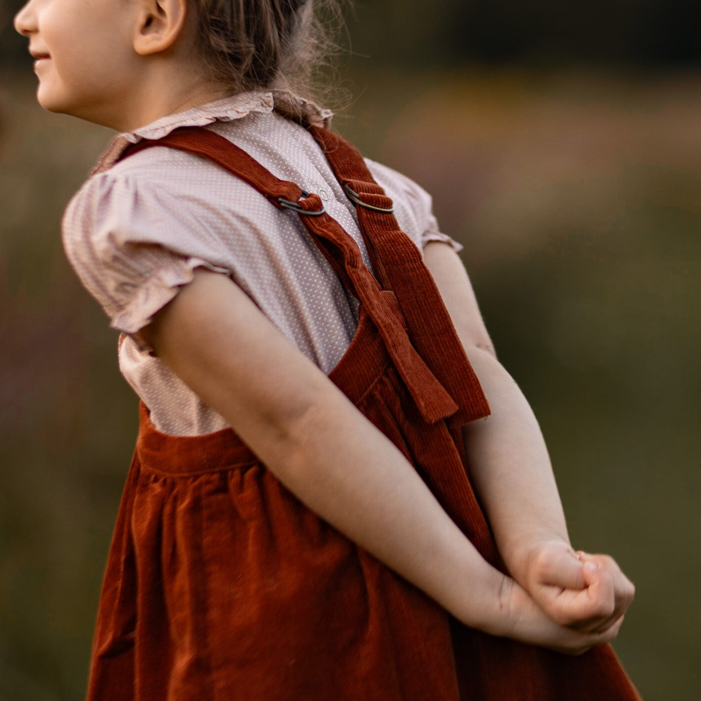 Child wearing a red dress with suspenders against a blurred natural background