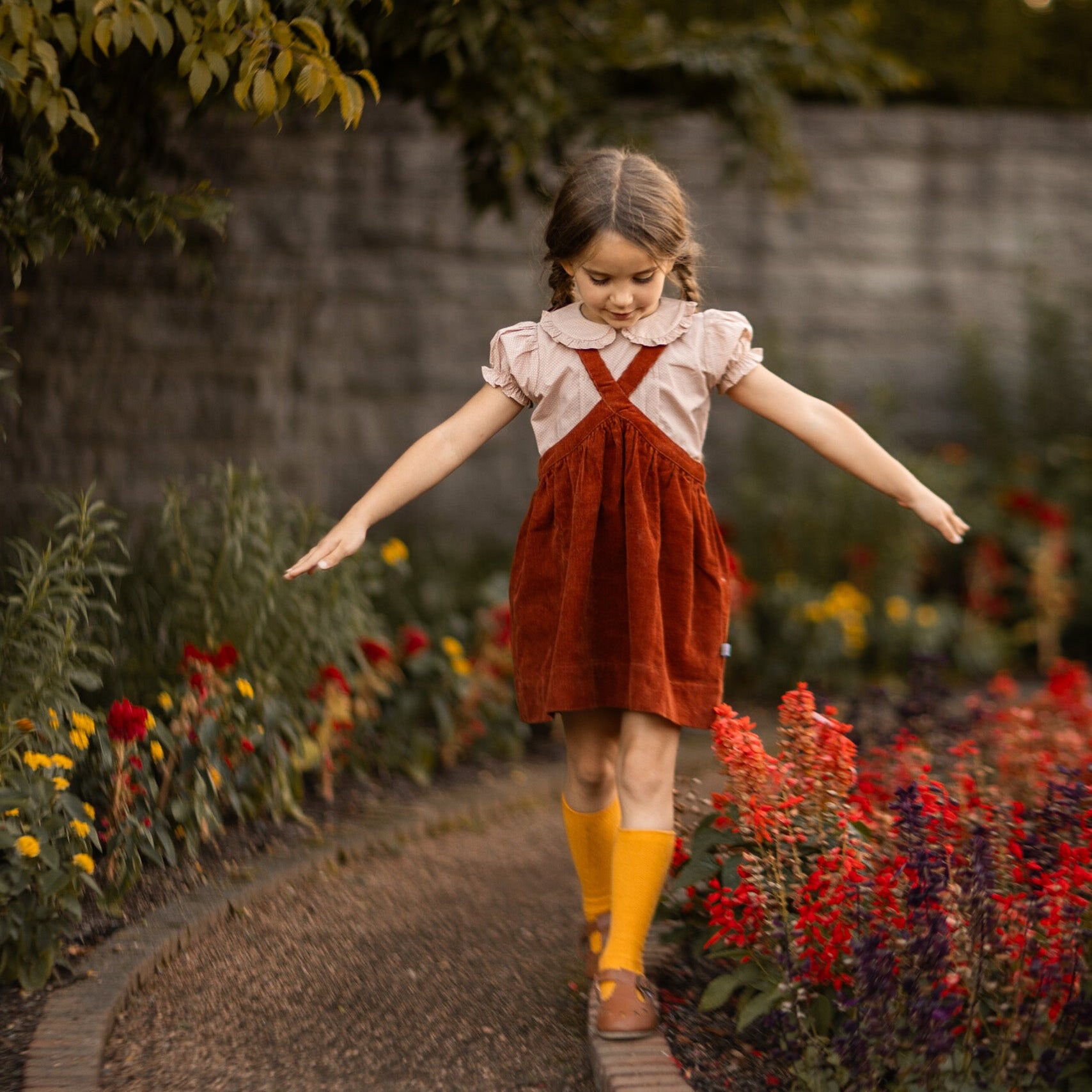 Young girl in a red dress and yellow socks standing on a garden path with flowers around.