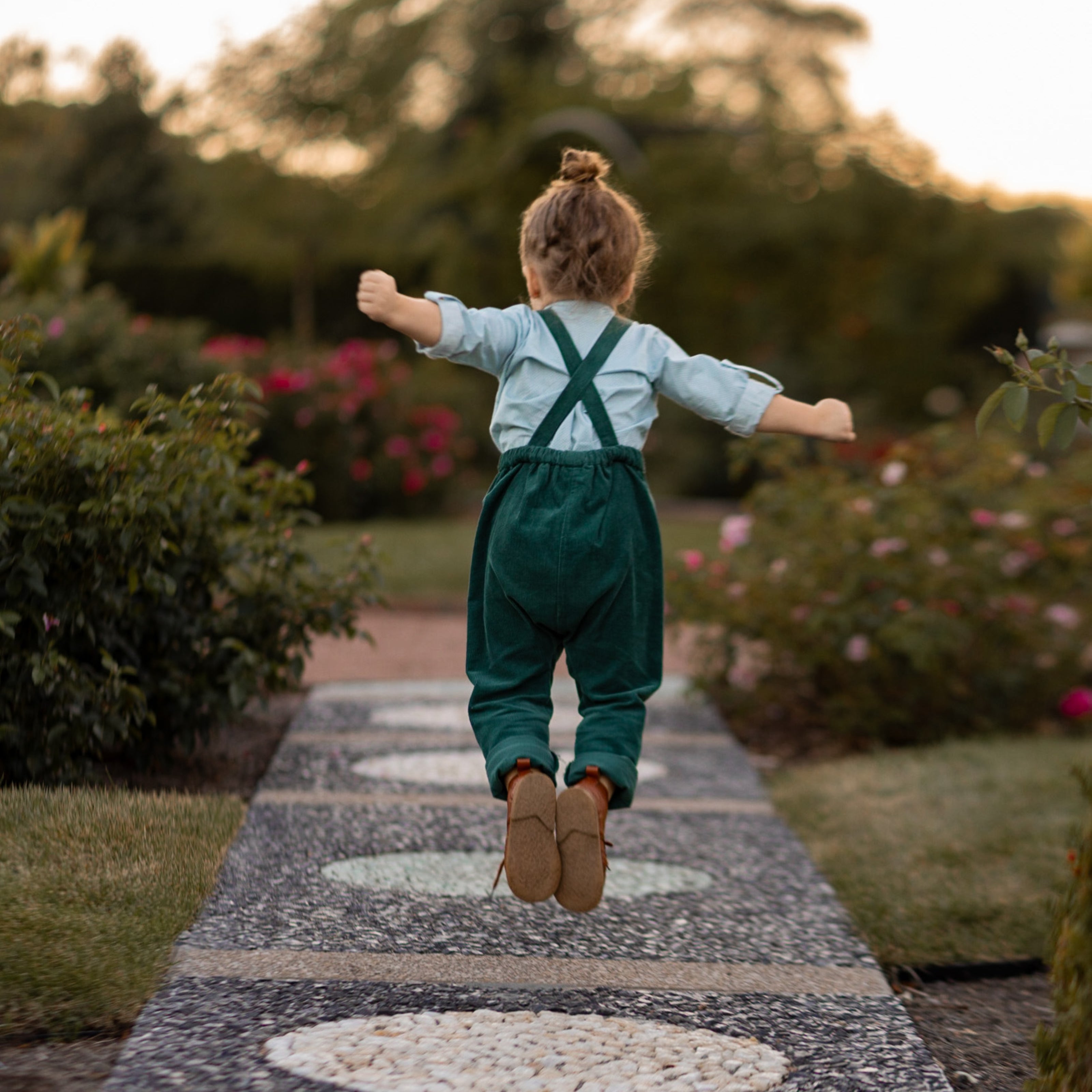 A 3-year-old model boy joyfully jumping to showcase the back of his classic light blue Henley shirt with white Swiss dots, made from natural, sustainable, and high-quality materials. The shirt features a timeless Henley placket, while the spruce corduroy long overalls add a rich texture and warmth. This dynamic pose highlights the outfit's playful style and comfort, perfect for active little ones.
