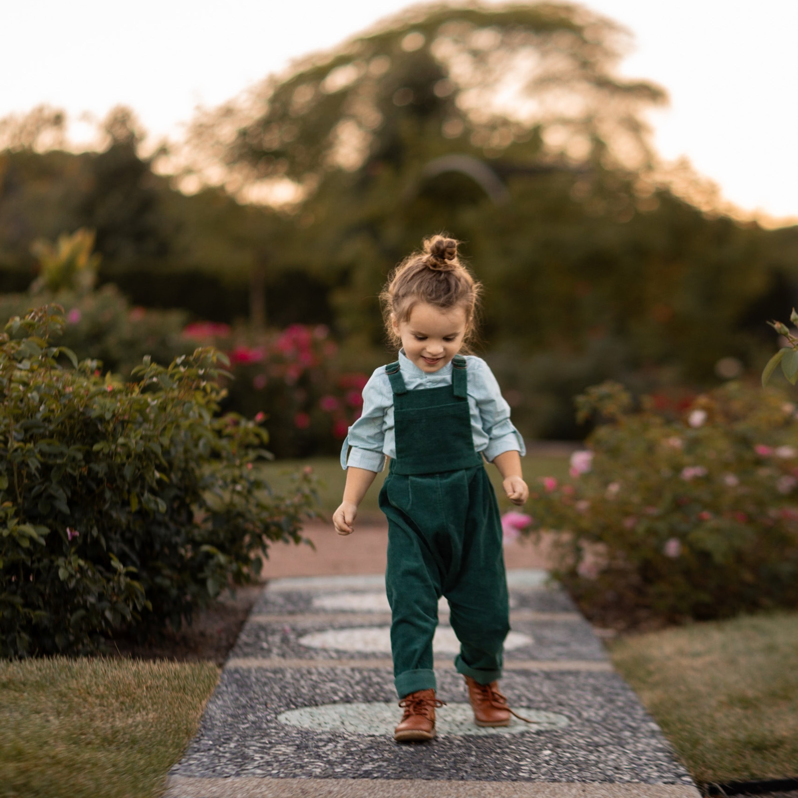 A 3-year-old model boy joyfully jumping outside in a classic light blue Henley shirt with white Swiss dots, made from natural, sustainable, and high-quality materials. He pairs it with spruce corduroy long overalls, which add texture and warmth. The playful action captures the spirit of childhood, showcasing the outfit's comfort and style for outdoor adventures.