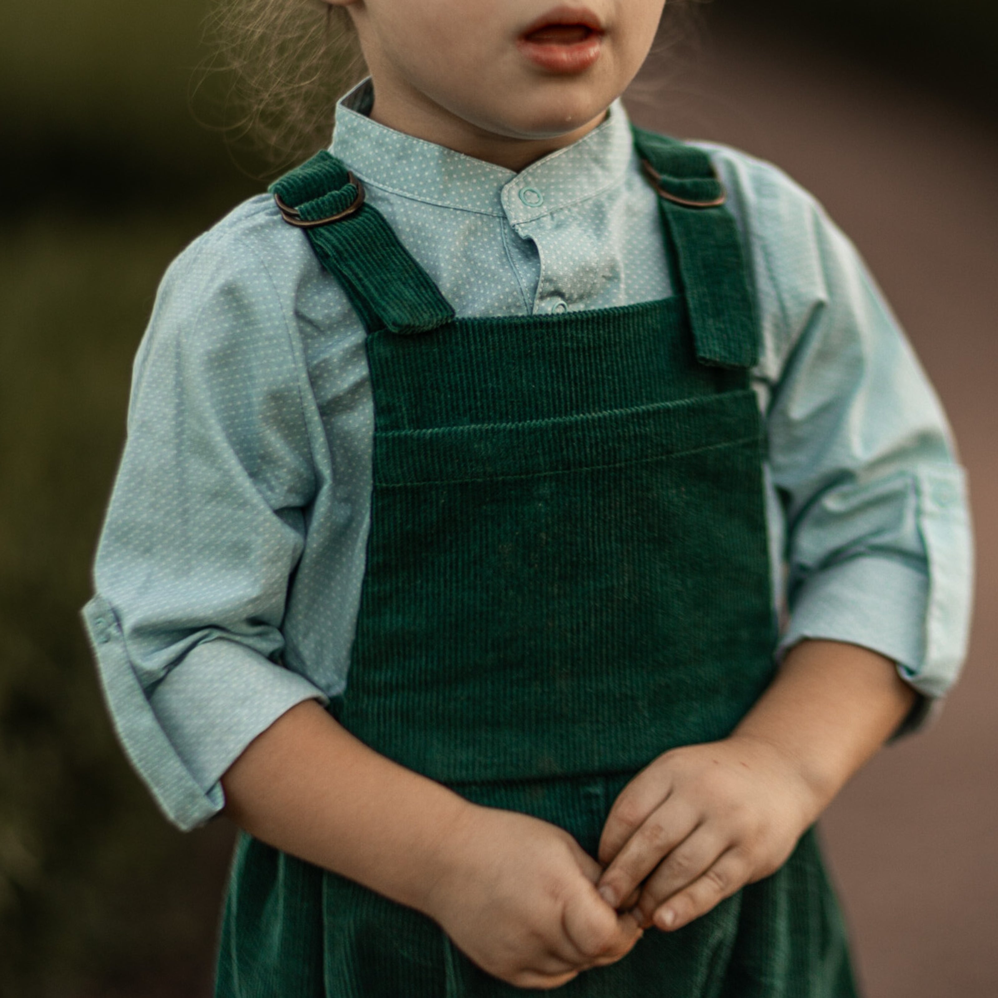 A 3-year-old model boy showcasing a classic light blue Henley shirt with white Swiss dots, crafted from natural, sustainable, and high-quality materials. He pairs it with spruce corduroy long overalls, which add a rich texture and warmth to the outfit. The combination highlights the playful yet stylish nature of children's fashion, perfect for both casual outings and special occasions.