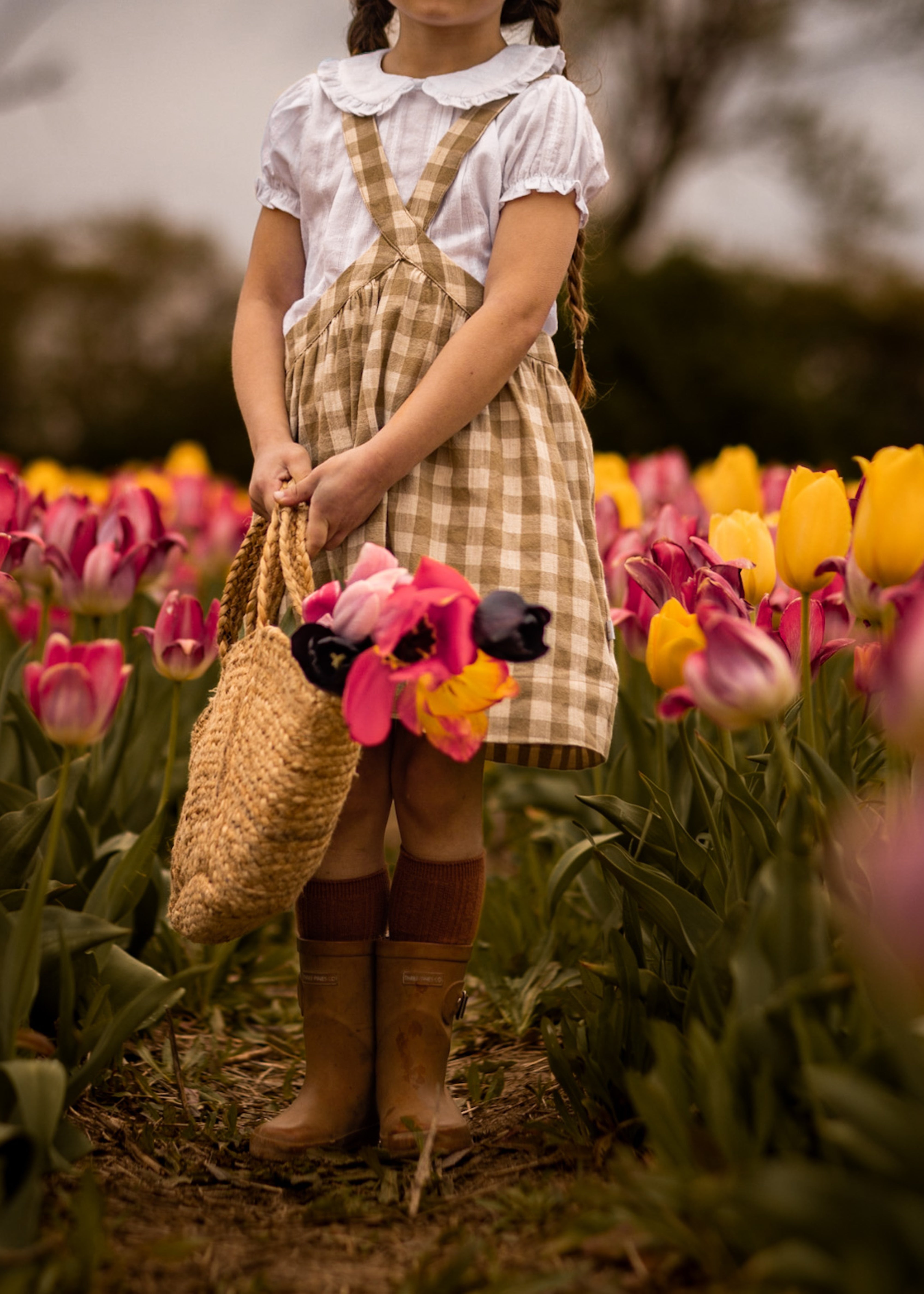 Child in a checkered dress standing in a tulip field holding flowers and a straw bag.