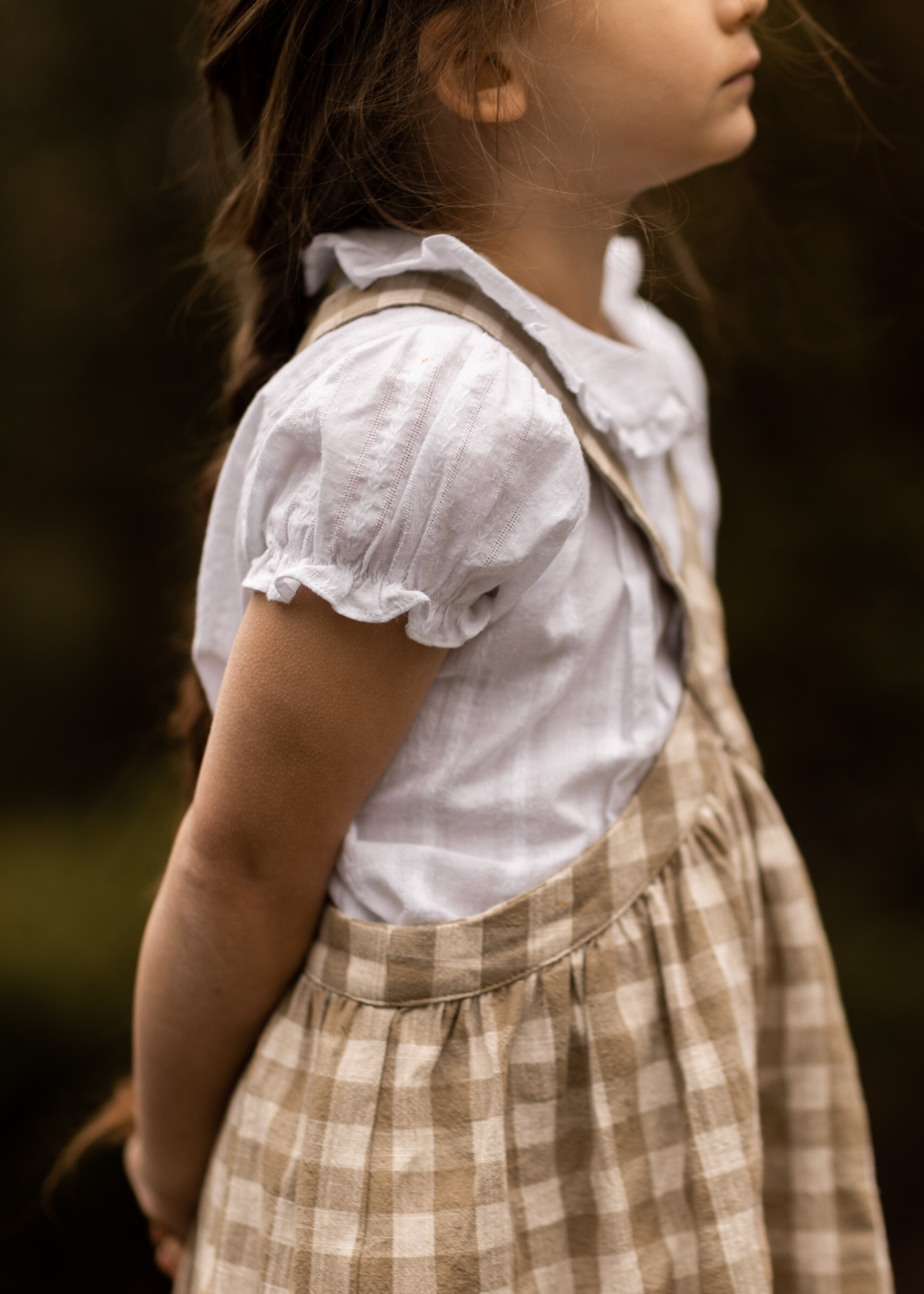 Young girl wearing a white blouse and brown checkered dress against a blurred background