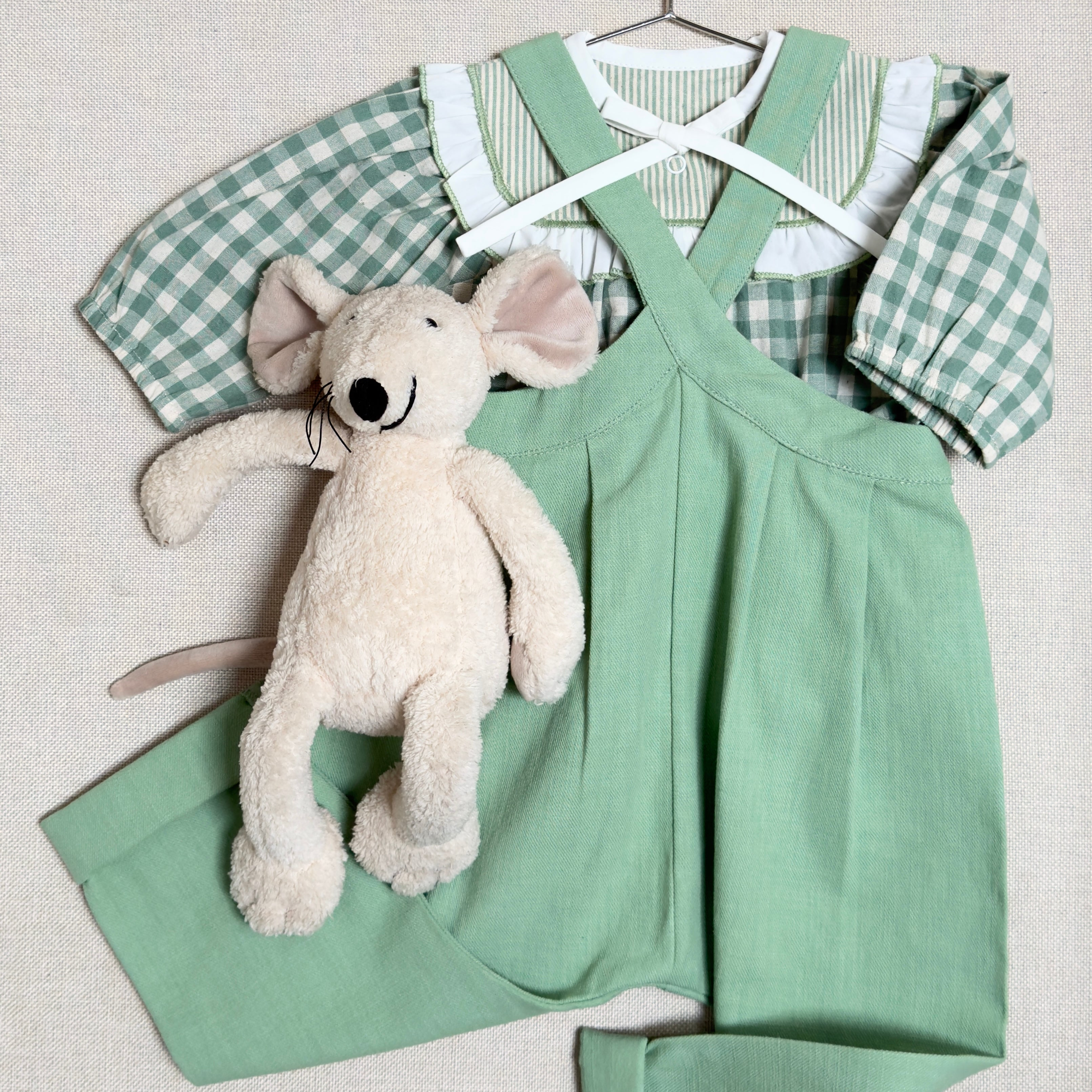 Green checkered shirt and green overalls with a plush toy on a hanger against a light background